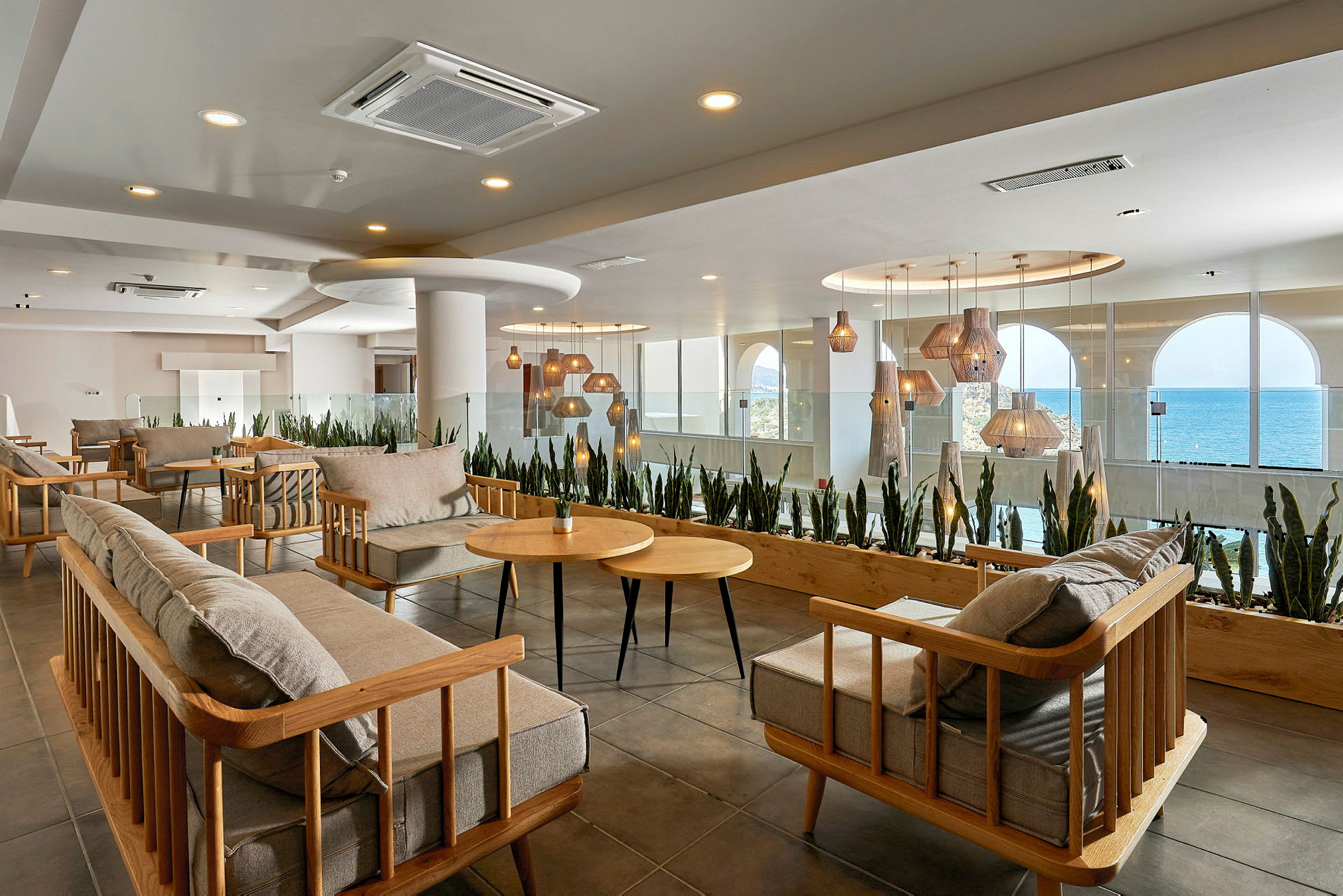 En moderne lounge med trestoler og bord samt hengende lamper. Store vinduer med utsikt over havet i bakgrunnen. (AI-generert tekst)