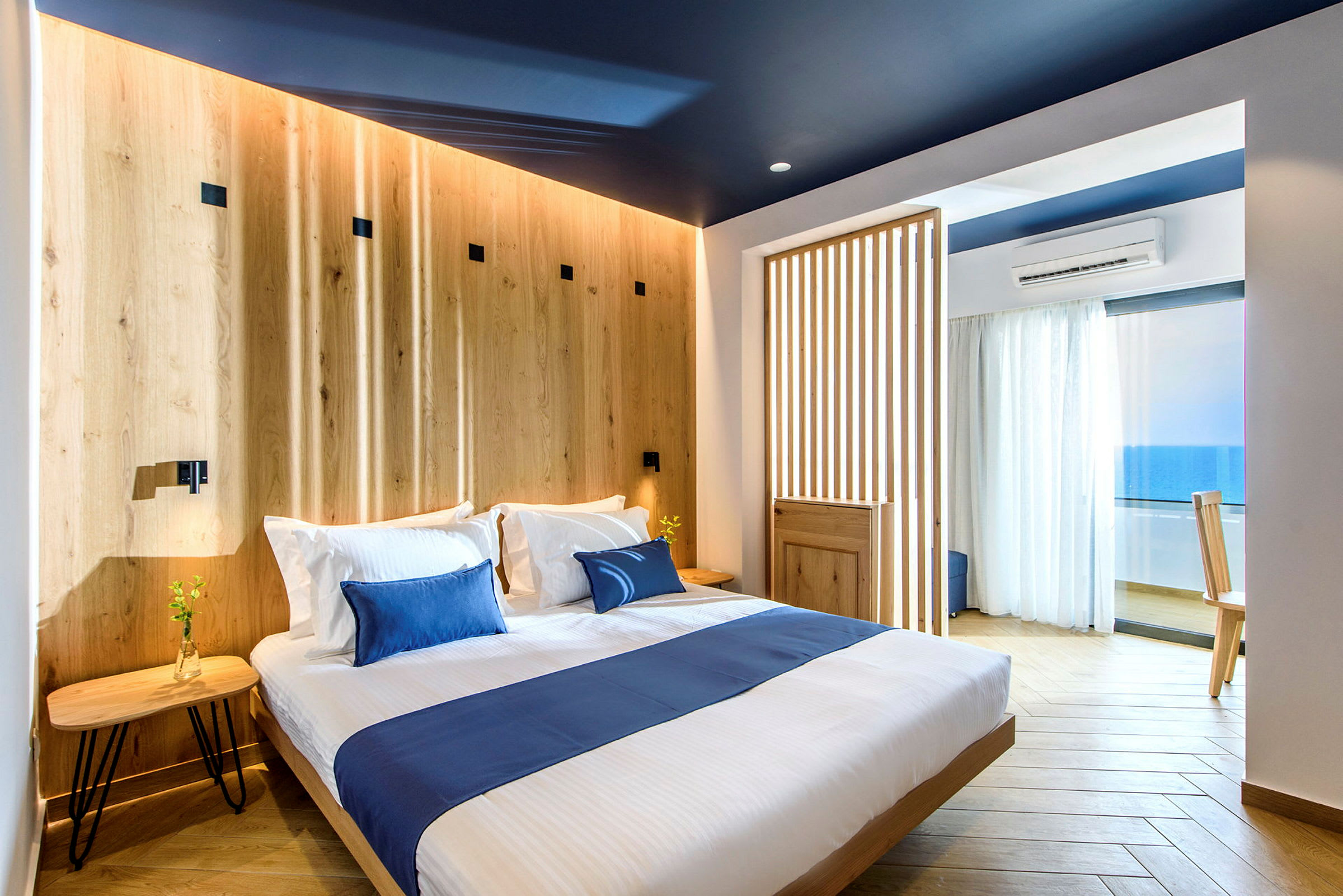 Et stilrent hotellrom med en dobbeltseng, innredet i lyst treverk og blå dekor, og utsikt over havet. (AI-generert tekst)