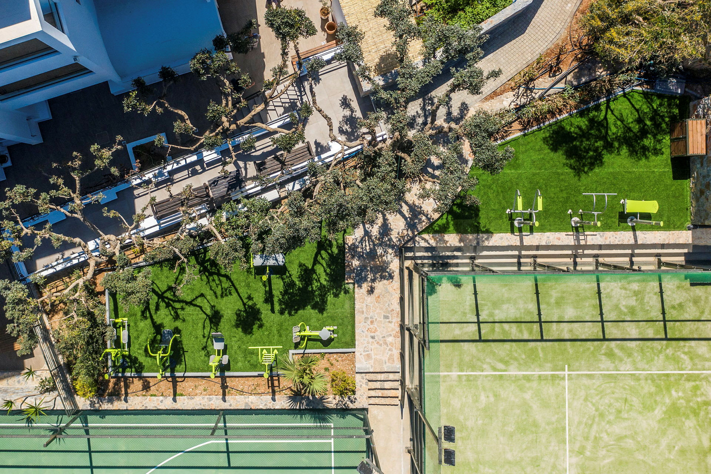 Flyfoto av en utendørsområde med trær, grønne plener, treningsutstyr og en tennisbane. (AI-generert tekst)