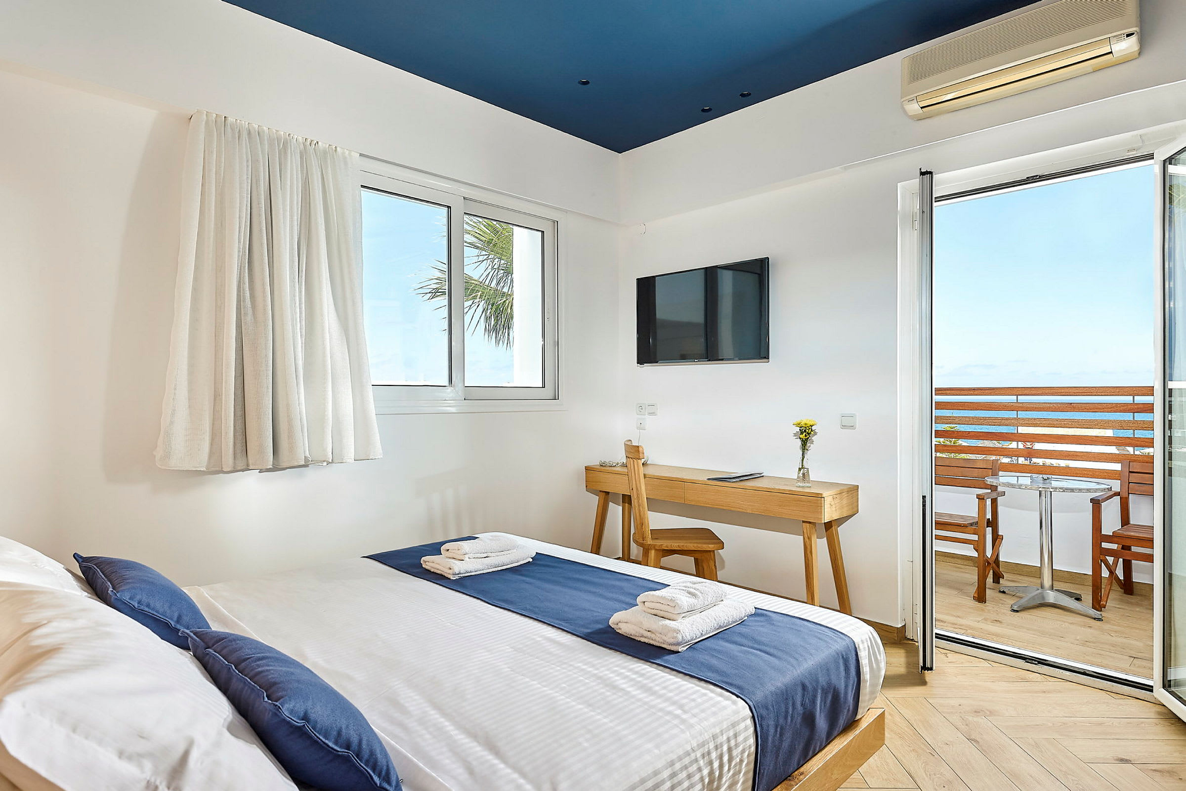Hotellrom med seng kledd i blått og hvitt sengetøy, skrivebord med stol, TV på veggen og balkong med utsikt over havet. (AI-generert tekst)