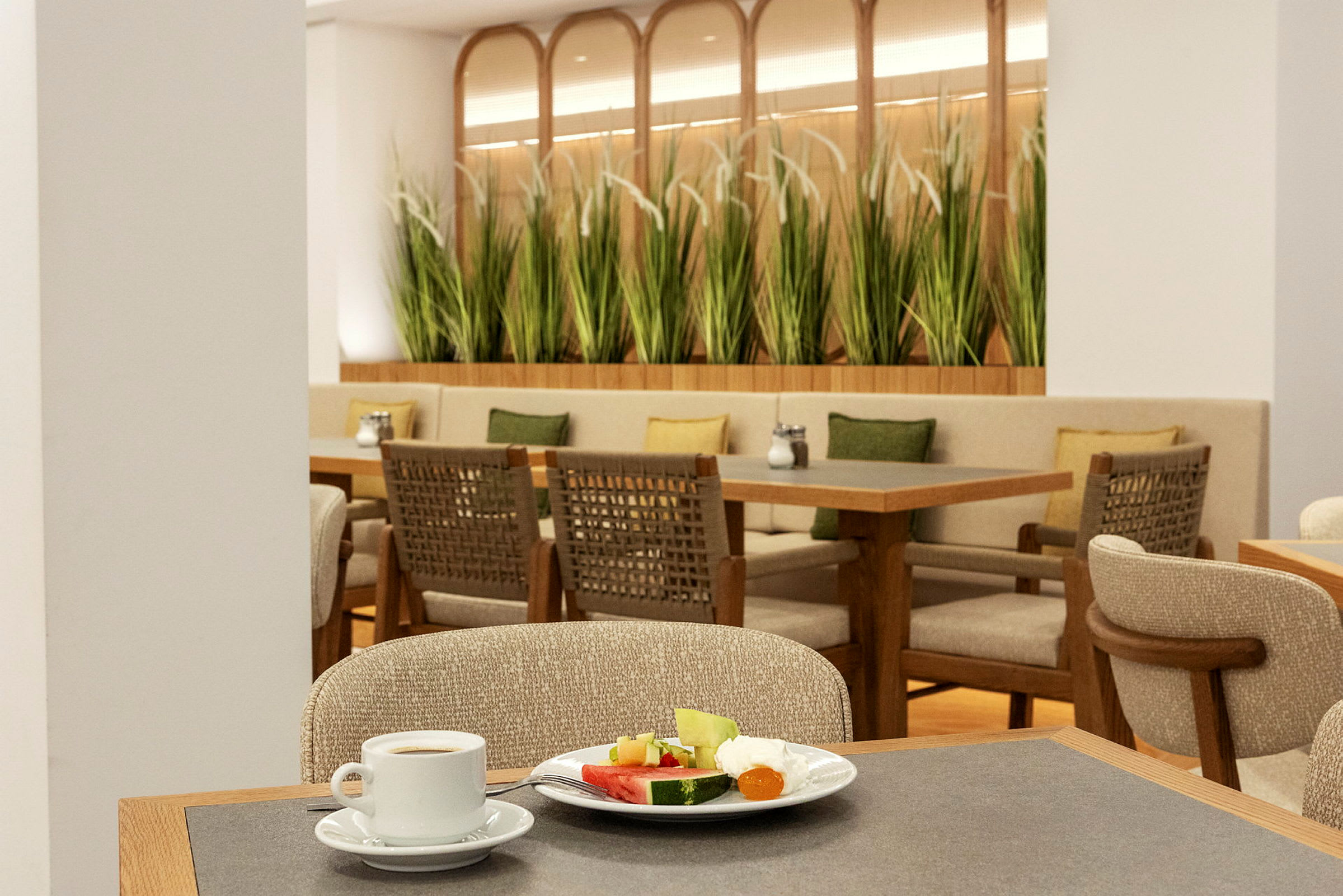 Bilde av en restaurant med bord, stoler og grønne planter som dekor. I forgrunnen sees en kaffekopp og en tallerken med frukt. (AI-generert tekst)