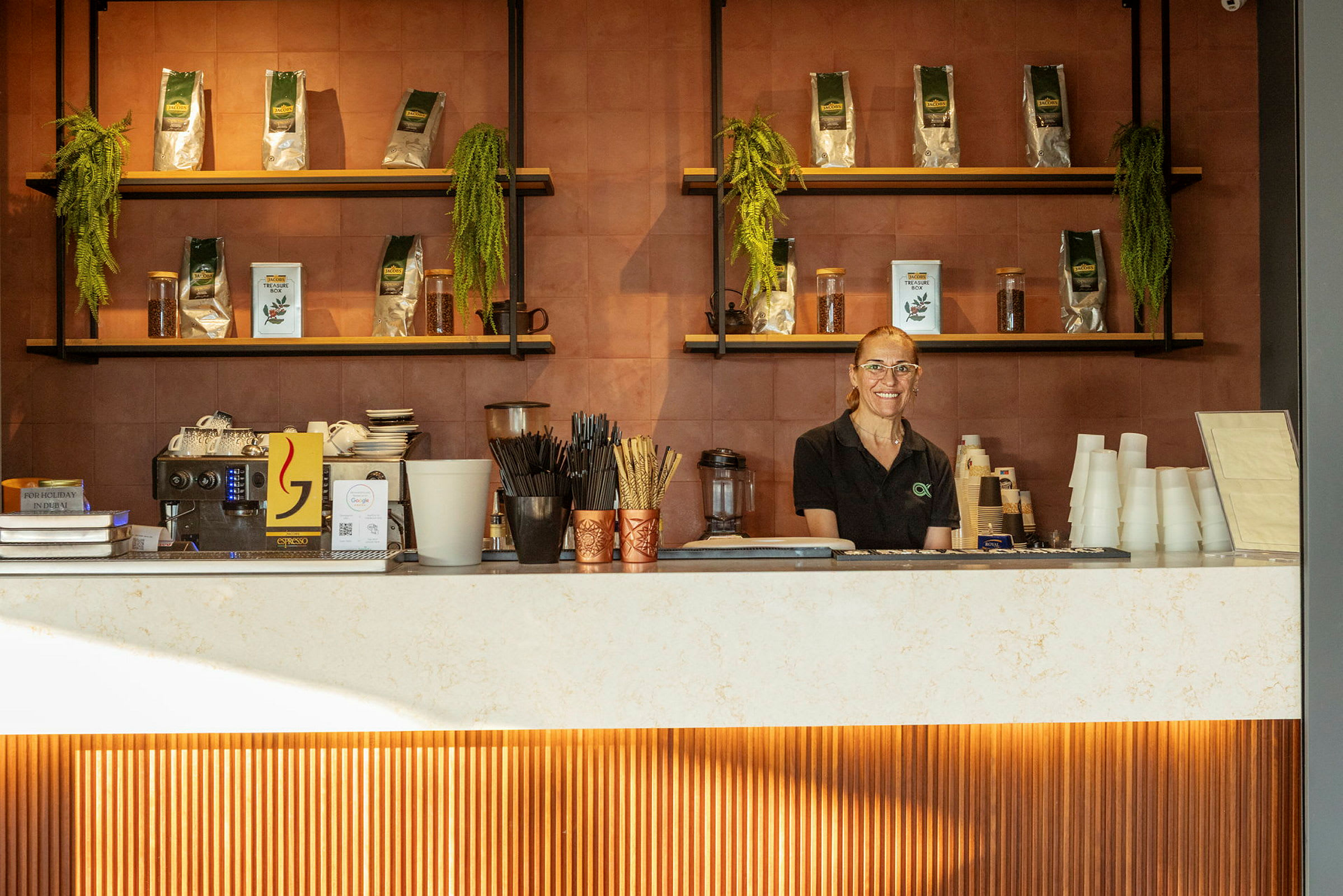 En person står bak disken i en kafé, omgitt av hyller med kaffeprodukter og dekorasjoner. (AI-generert tekst)