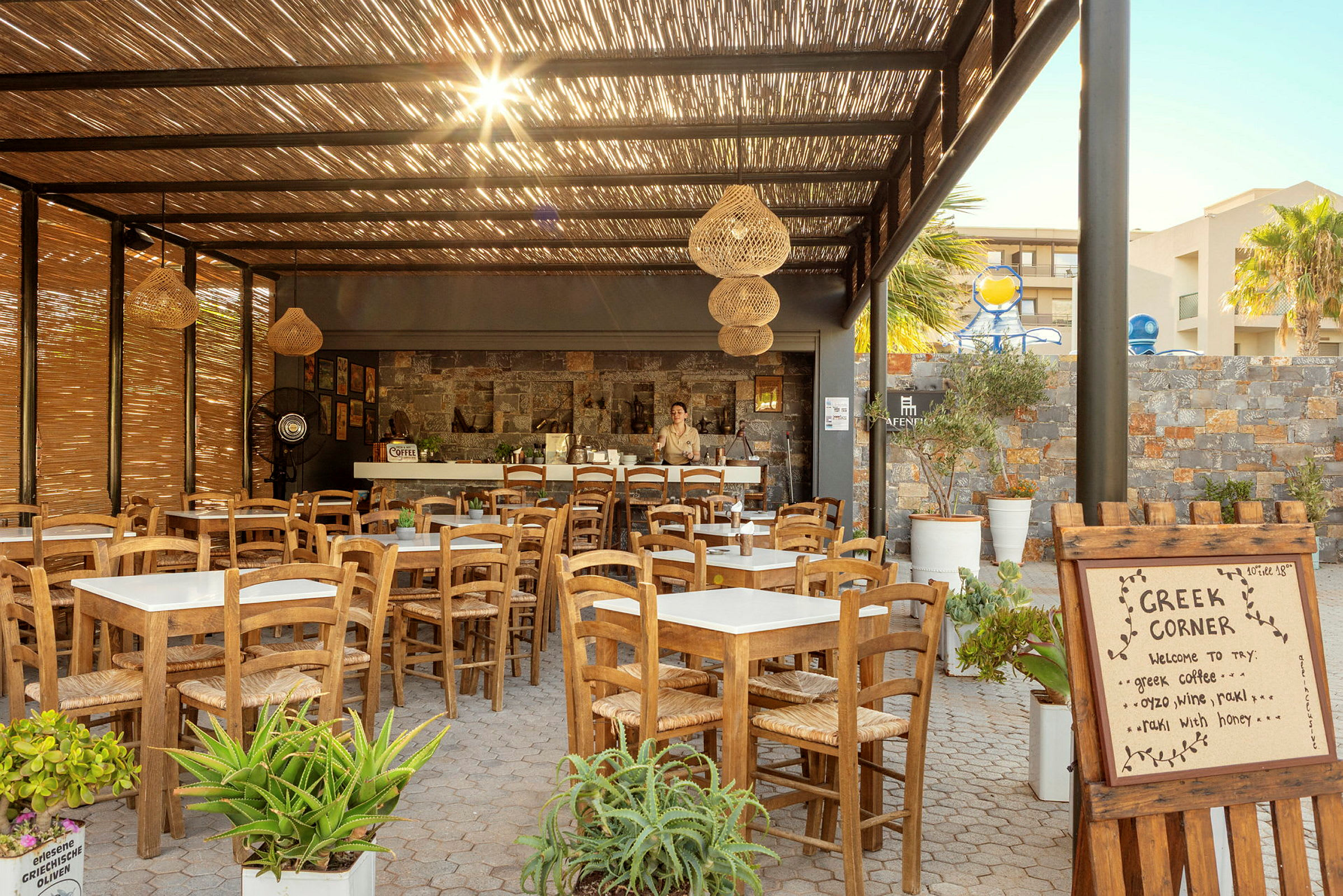 Bildet viser en utendørs restaurant med trebord og stoler under et tak av bambus, samt et skilt med teksten "Greek Corner". (AI-generert tekst)
