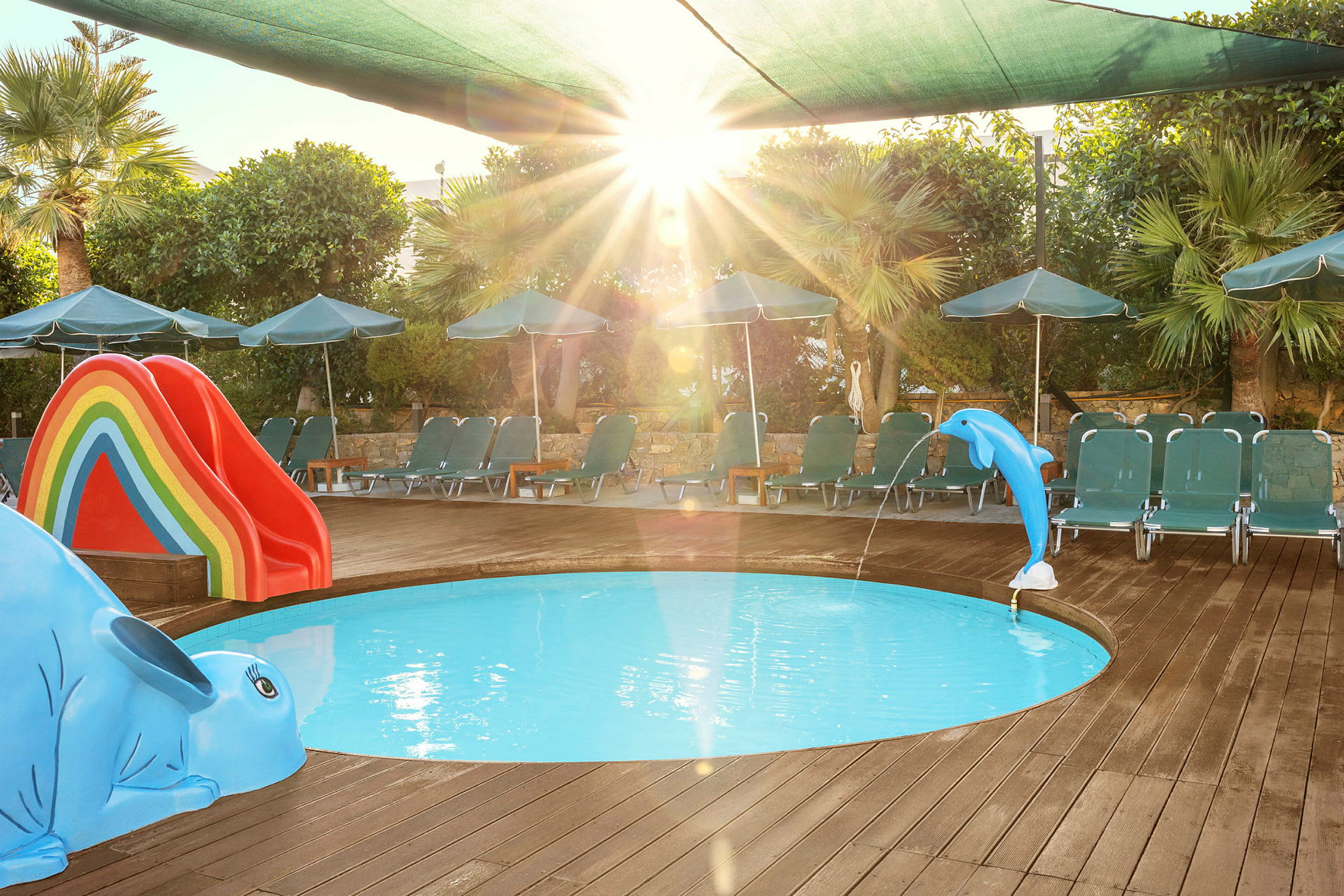 Et lite rundt barnebasseng omgitt av en treterrasse. I bassenget er en blå delfinfigur som spruter vann og en sklie i regnbuens farger. Solsenger og parasoller står rundt bassenget. (AI-generert tekst)
