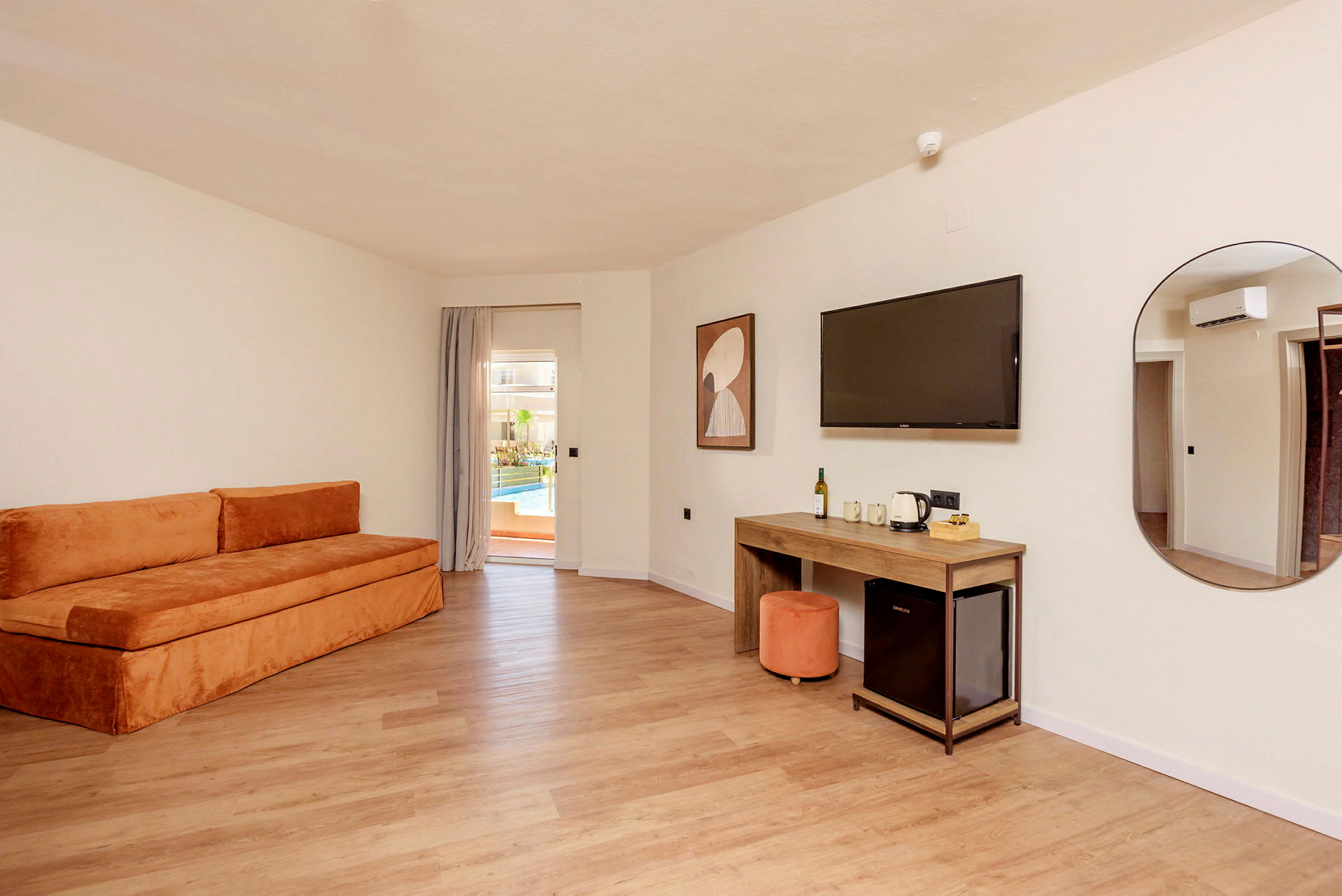 Et hotellrom med en oransje sofa, et skrivebord, en veggmontert TV og en dør til en terrasse med basseng. (AI-generert tekst)