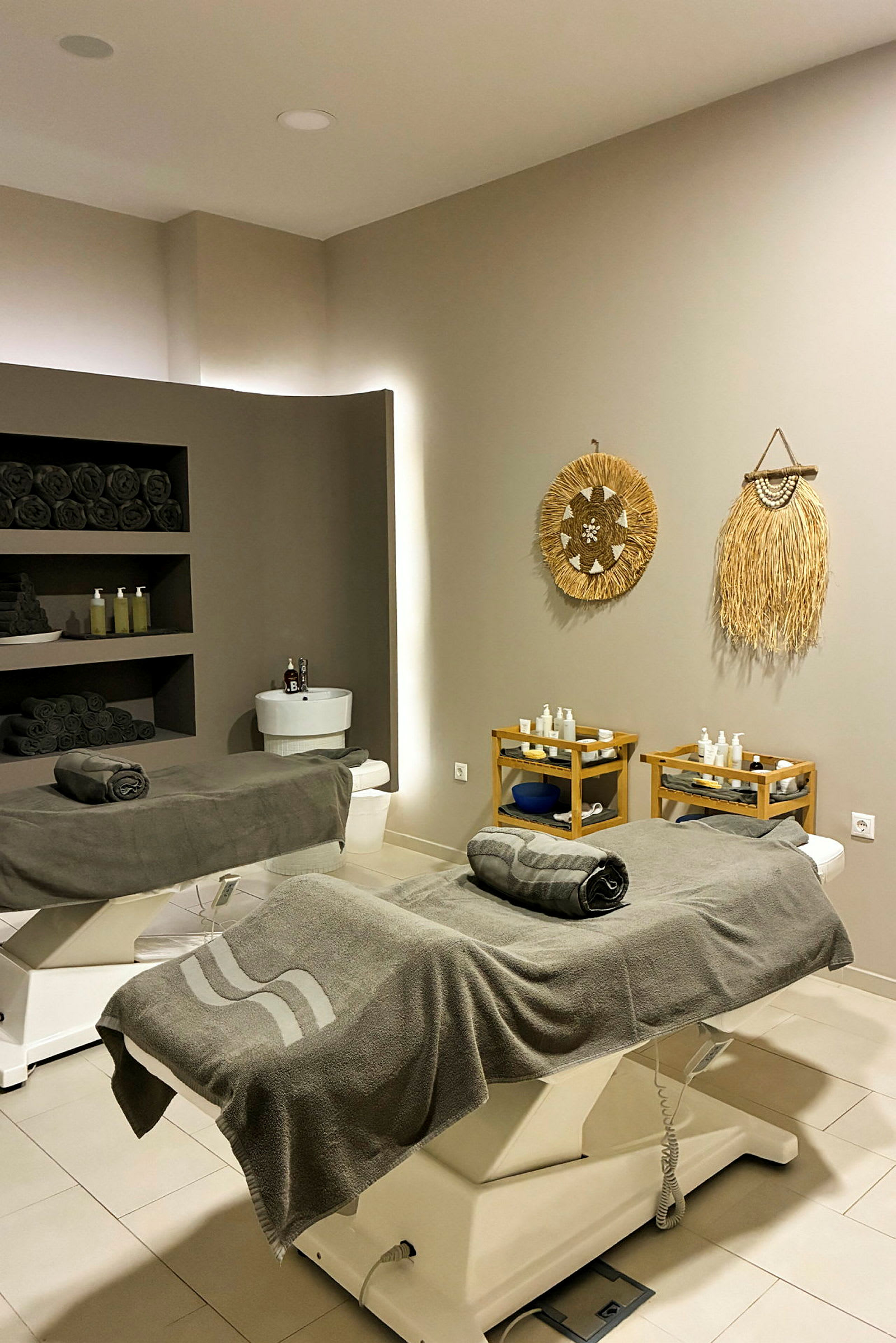 Et spa-rom med to behandlingsbenker kledd i grått frottéhåndklær. Veggene er dekorert med naturinspirerte kunstverk, og hyller med mørke håndklær og produkter er synlige i bakgrunnen. (AI-generert tekst)