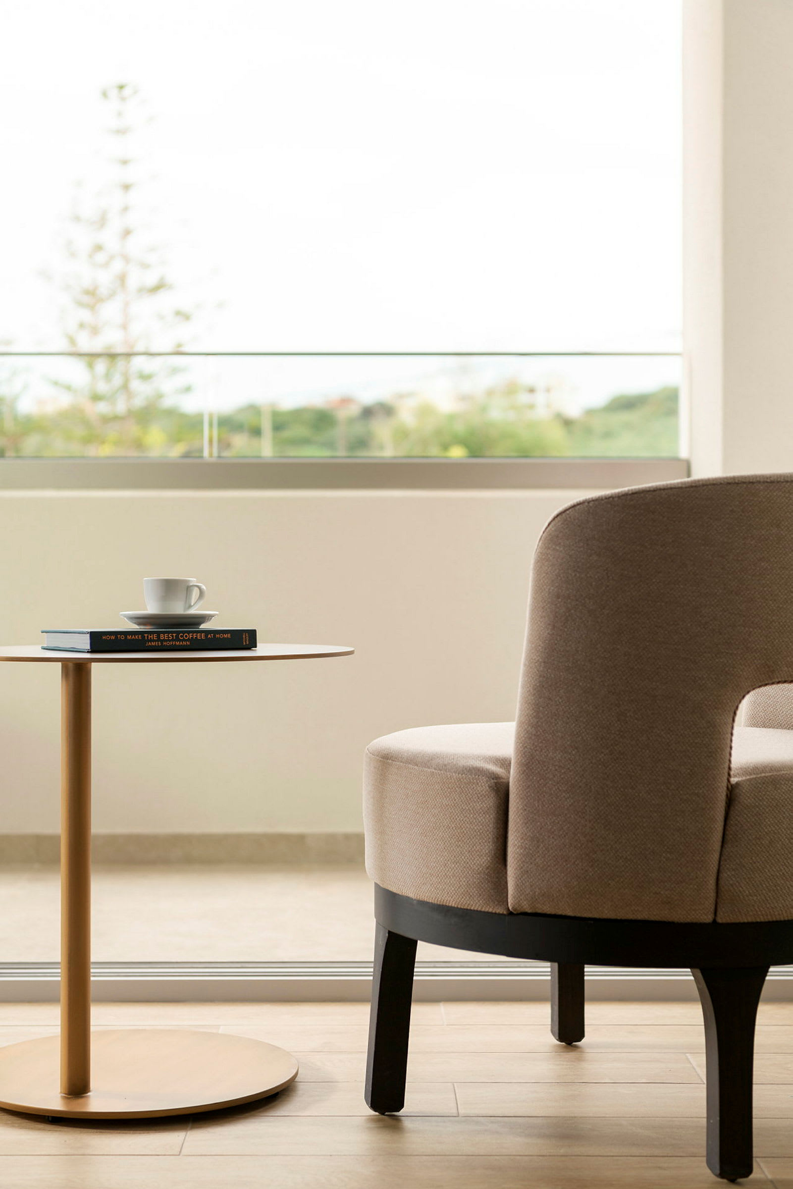 Interiørbilde som viser en beige lenestol og et lite sidebord med en bok og en kaffekopp. I bakgrunnen er et åpent vindu med grønn utsikt. (AI-generert tekst)