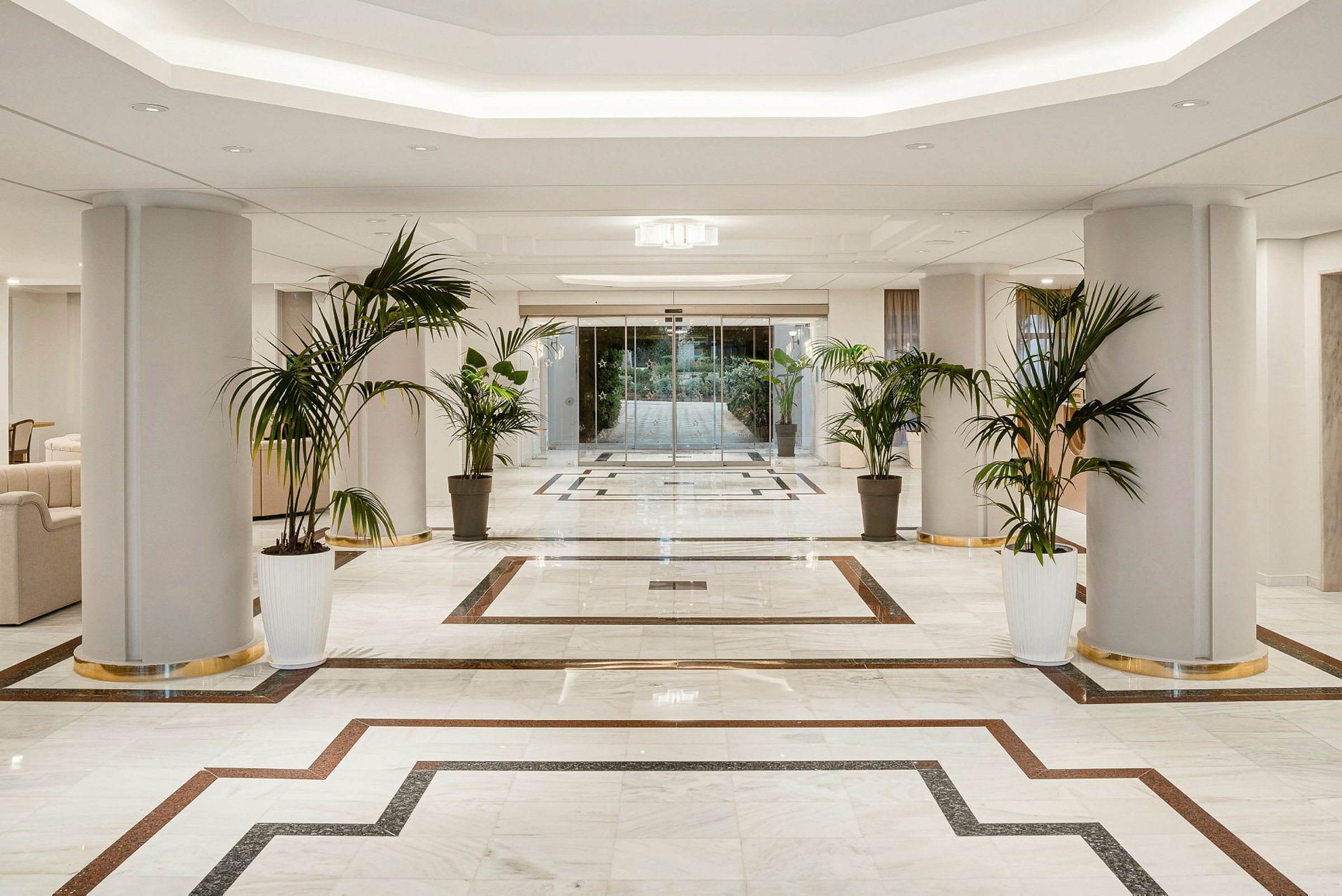 Lys og elegant hotellobby med marmorgulv, søyler og store grønne planter plassert i krukker. (AI-generert tekst)