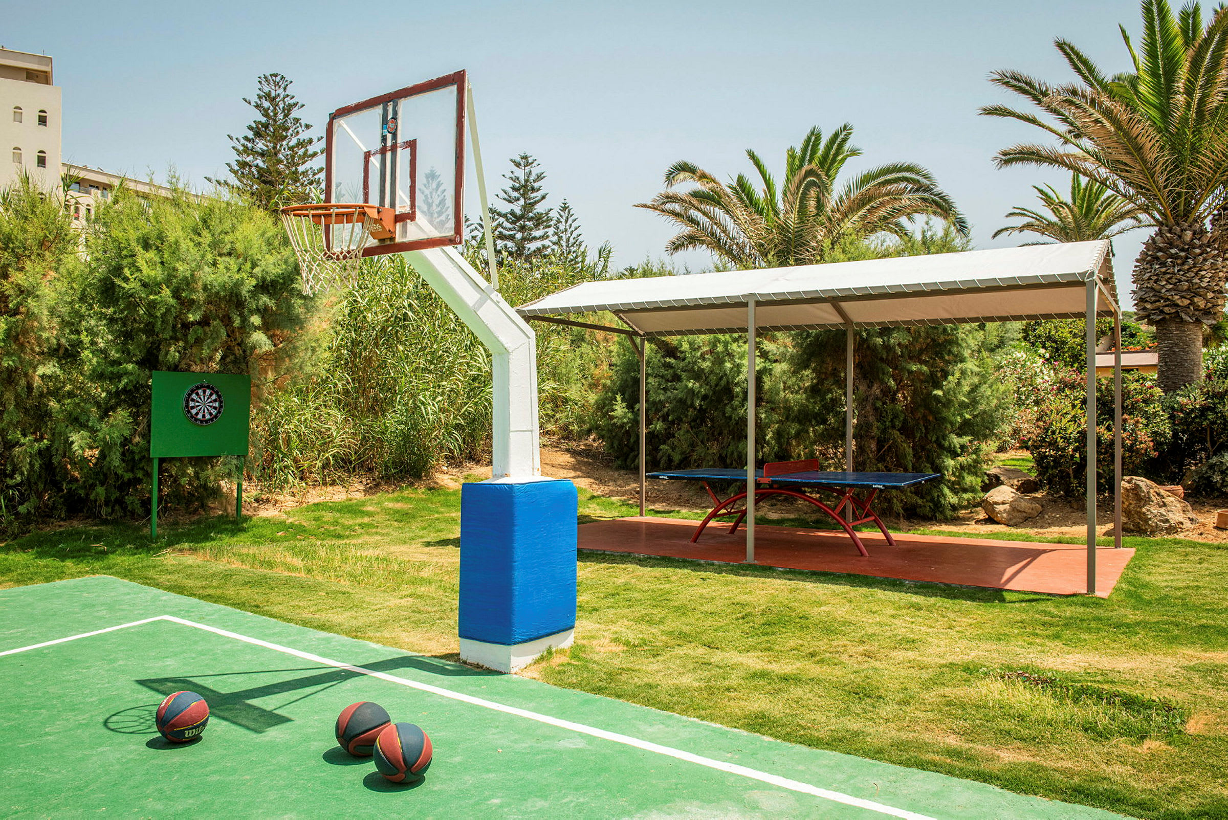En utendørs basketballbane med tre basketballer på bakken foran et basketballnett. En darttavle er også synlig til venstre, og i bakgrunnen er det et bordtennisbord under en overdekket struktur. (AI-generert tekst)