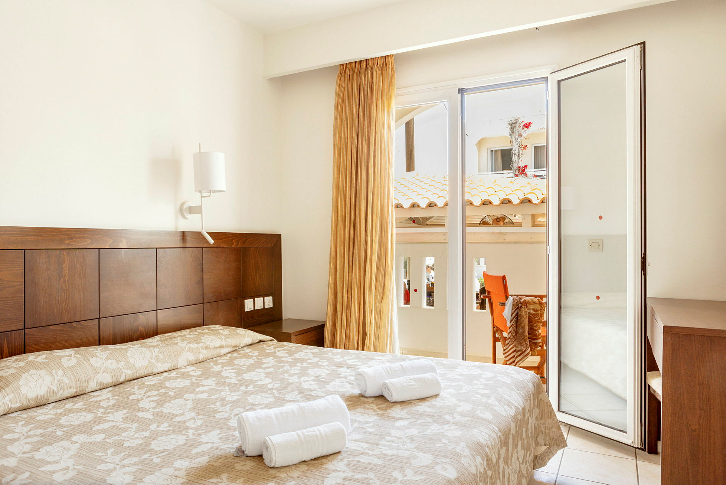 Et hotellrom med en dobbeltseng, lyst sengeteppe og håndklær oppå. Ved siden av er det en glassdør som fører ut til en balkong med utsikt over bygninger. (AI-generert tekst)