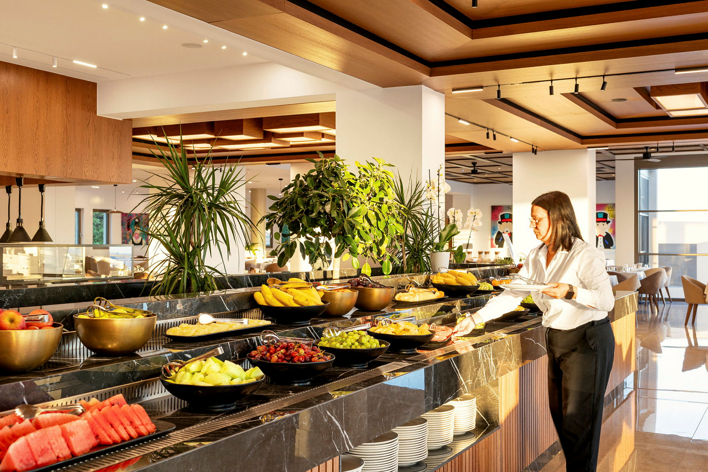En frokostbuffet med ulike valg av frukt og grønnsaker pent arrangert i skåler på et bord. En person står ved siden og tar mat. (AI-generert tekst)
