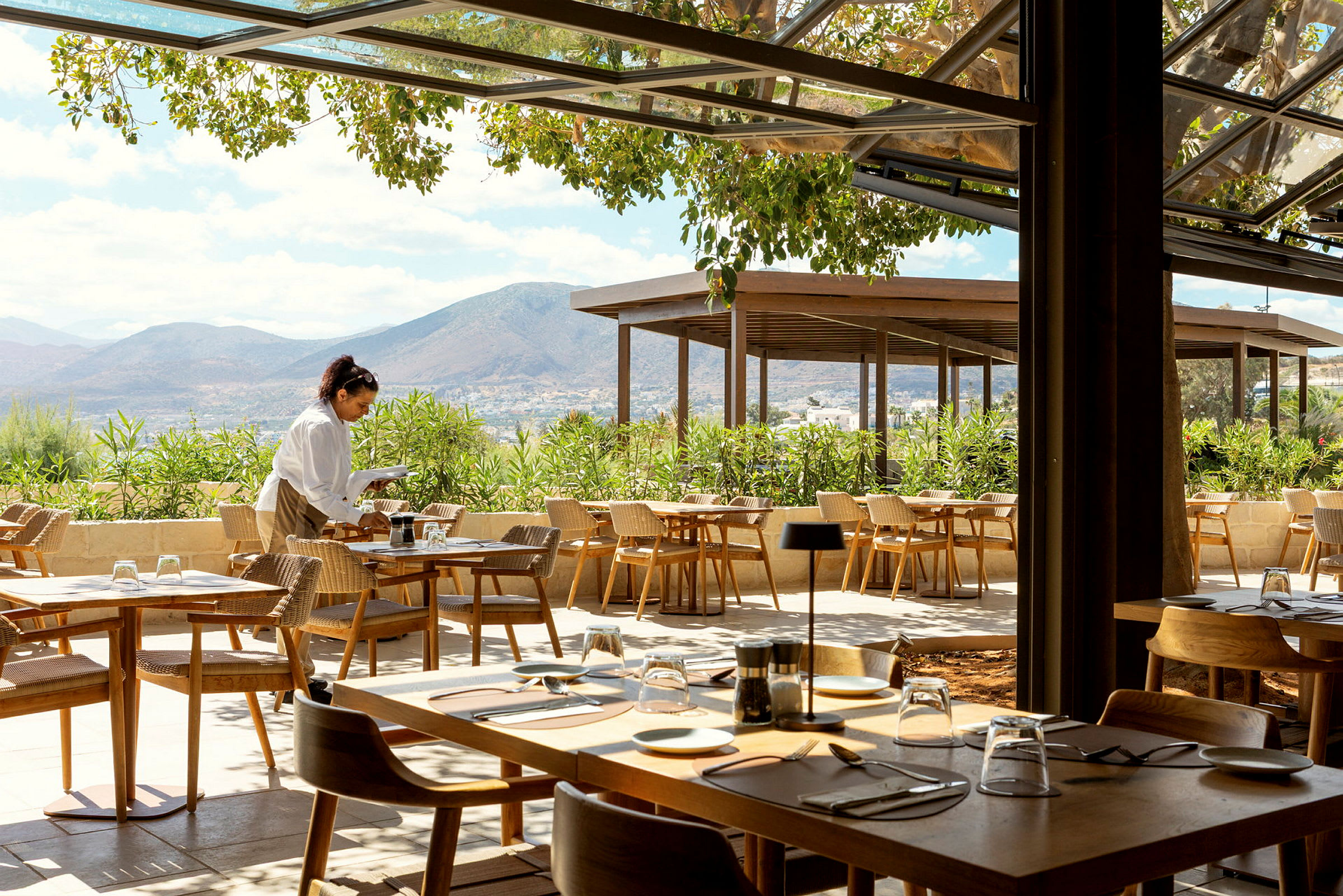 En utendørsrestaurant med trebord og stoler, omgitt av grøntområder og med utsikt over fjellene i bakgrunnen. En servitør går mellom bordene. (AI-generert tekst)