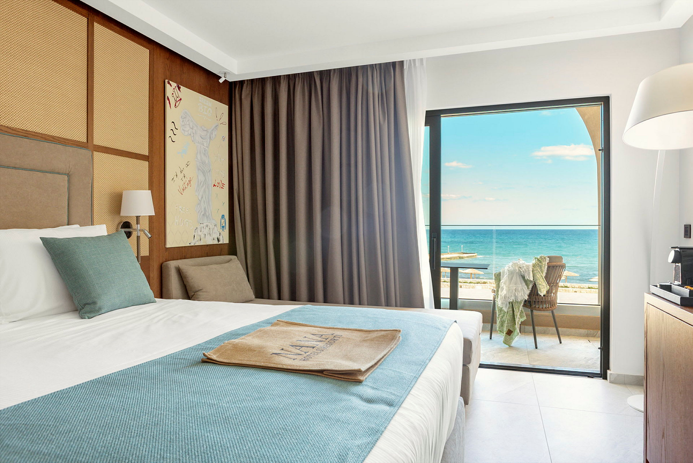 Hotellrom med en oppredd seng med hvitt og blått sengetøy. Rommet har store vinduer med utsikt over havet og en møblert balkong. (AI-generert tekst)