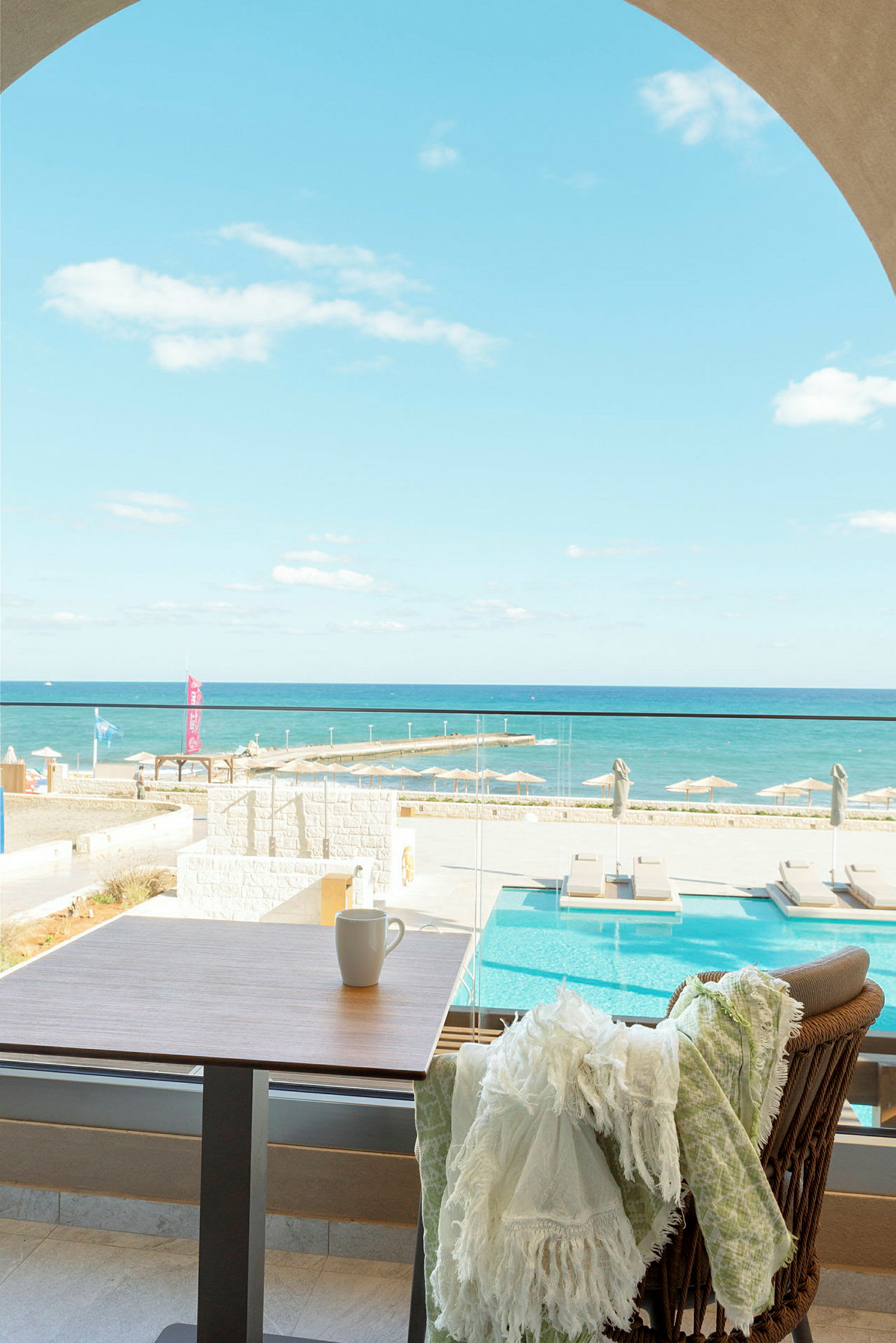 Utsikt over et basseng og havet fra en terrasse, med et bord og en stol i forgrunnen. (AI-generert tekst)