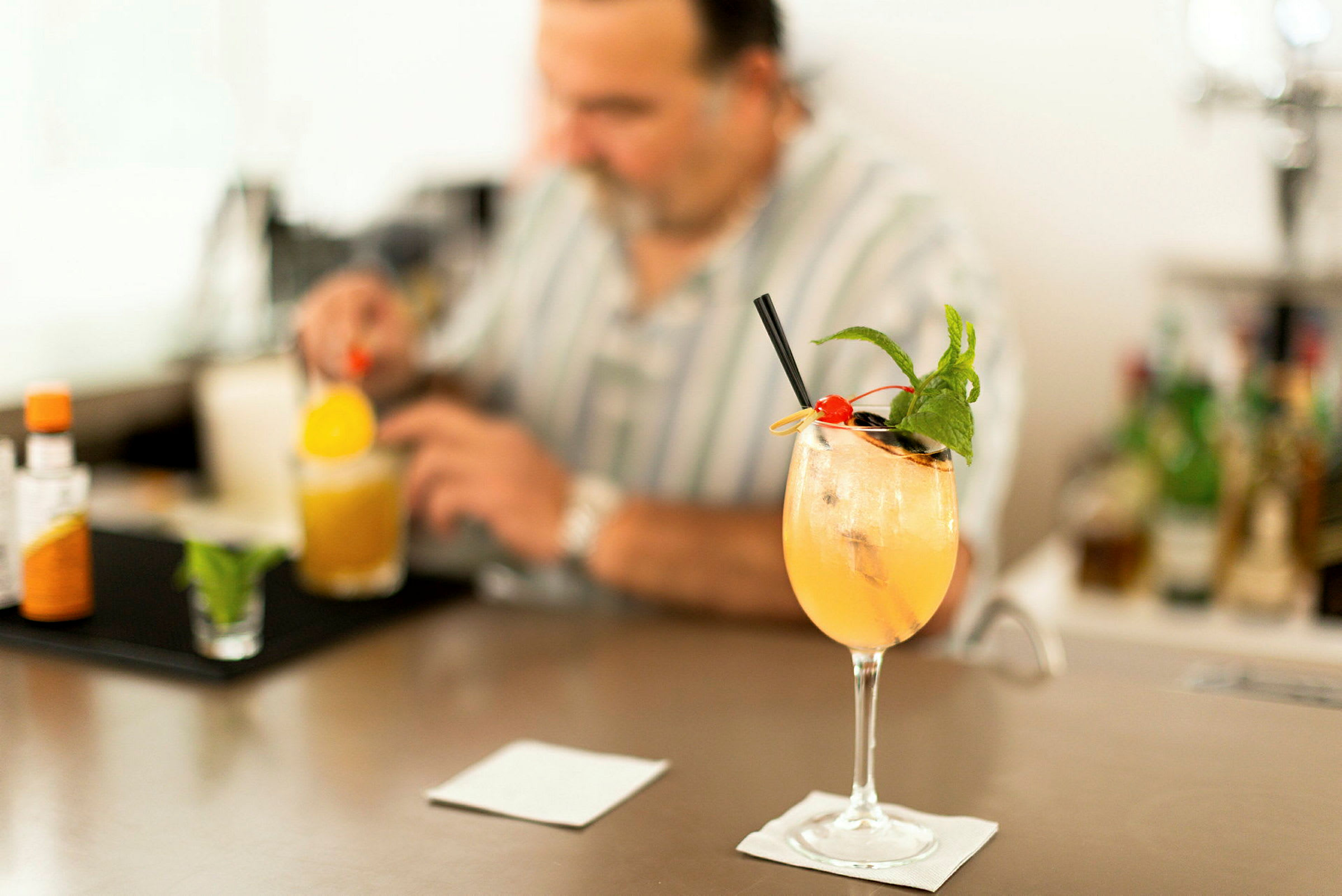 Et cocktailglass med en fargerik drikk, toppet med is, mynte og et kirsebær, står på en bardisk. En person i bakgrunnen blander en drink. (AI-generert tekst)