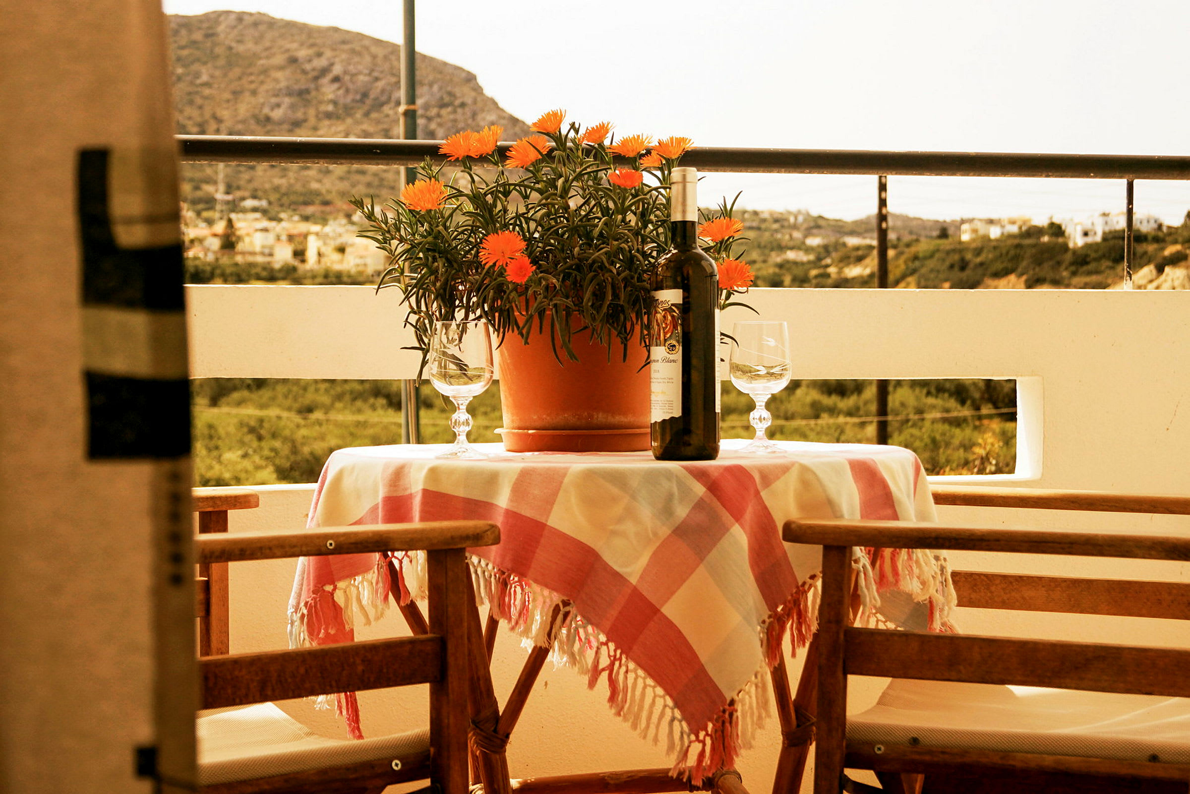 En terrasse med et bord dekket med en rutete duk, en potteplante, en vinflaske og to glass. Bakgrunnen viser en naturskjønn utsikt. (AI-generert tekst)