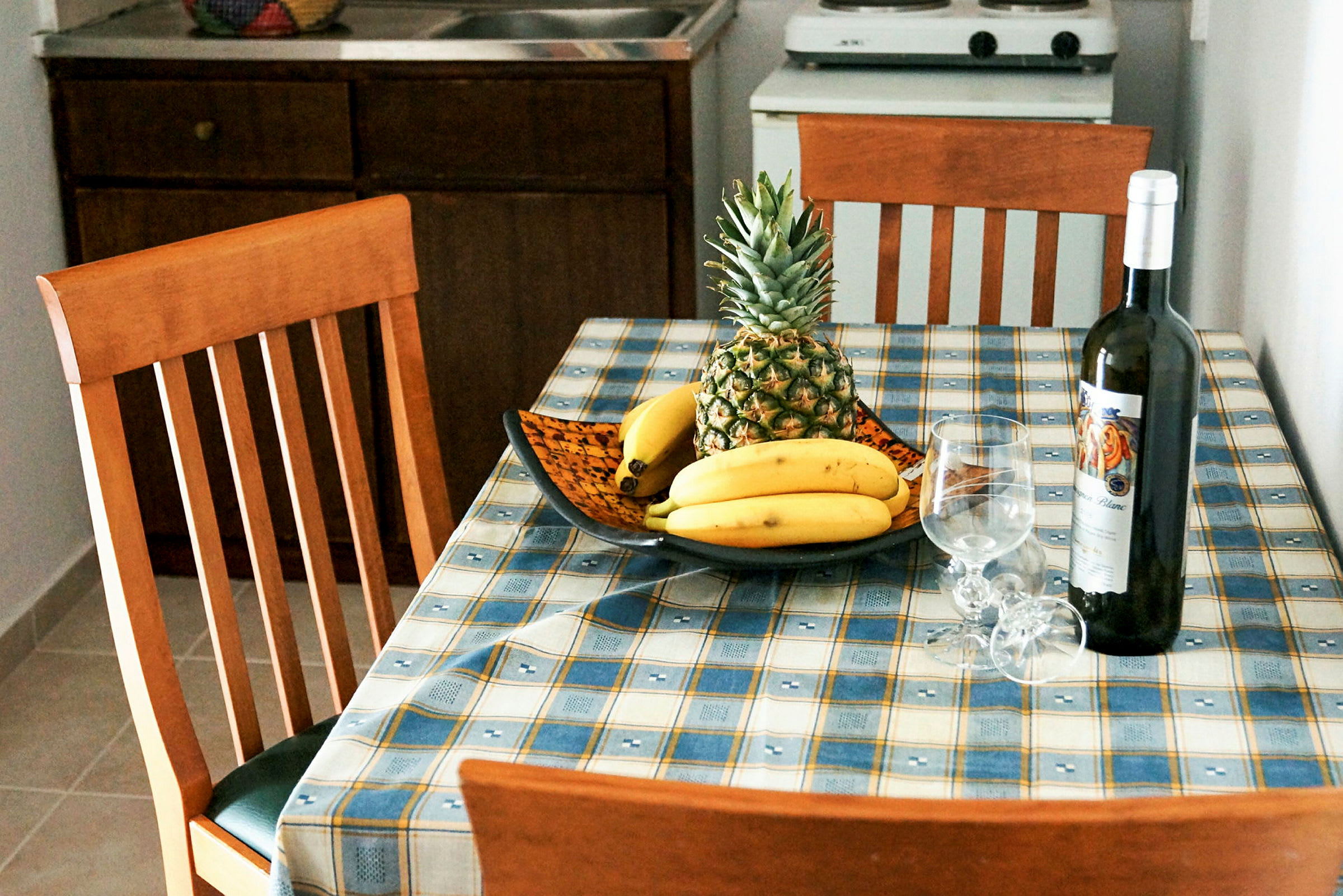 Et kjøkkenbord med en fruktskål med ananas og bananer, samt en vinflaske og to vinglass. (AI-generert tekst)
