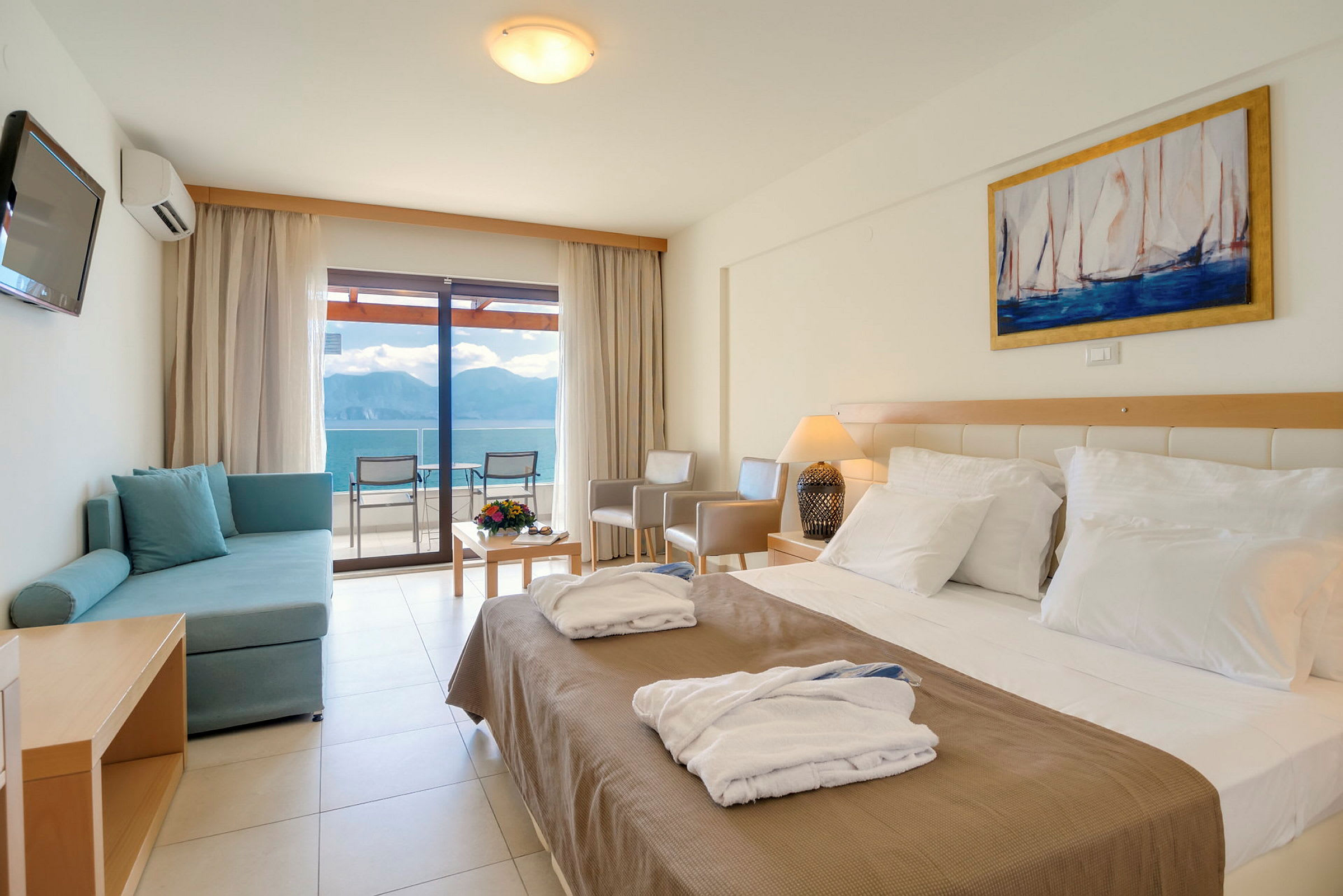 Et hotellrom med en dobbeltseng, en sofa, et bord med blomster og utsikt over havet gjennom glassdører. (AI-generert tekst)