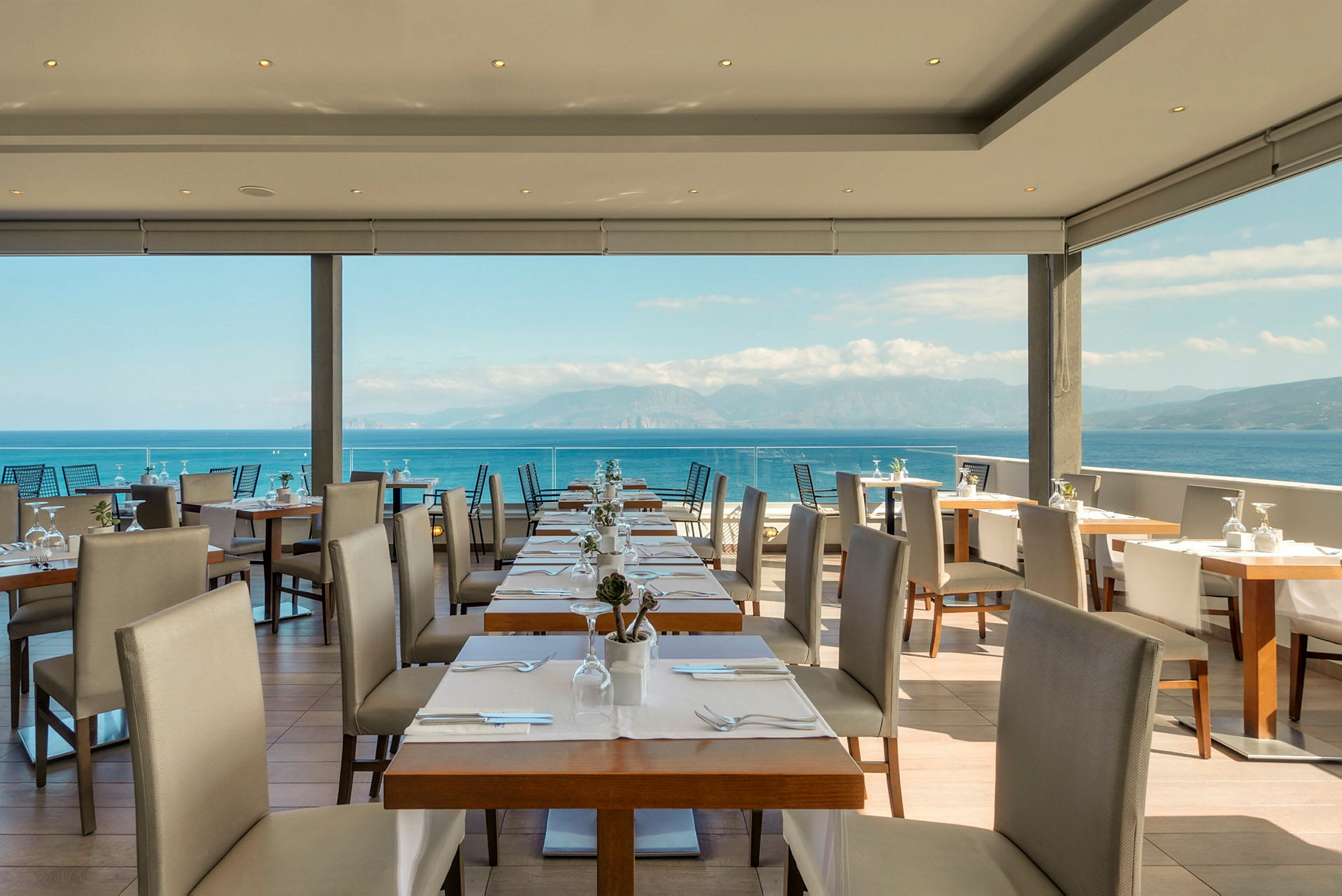 Utsikt over en restaurant med dekkede bord og stoler, med havet og fjellene i bakgrunnen. (AI-generert tekst)