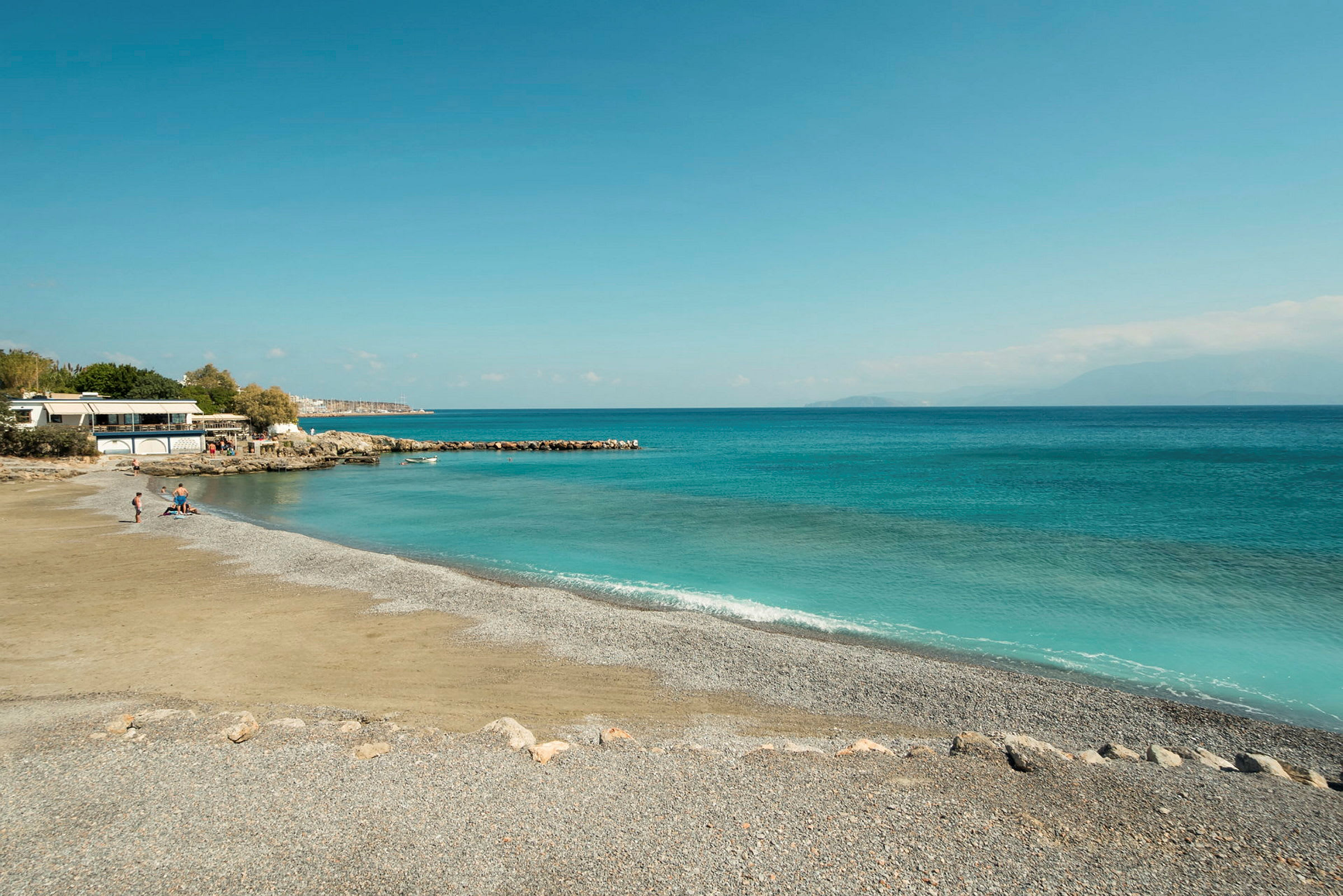 Strand med småstein og klart blått vann, med noen mennesker nær en bygning. (AI-generert tekst)