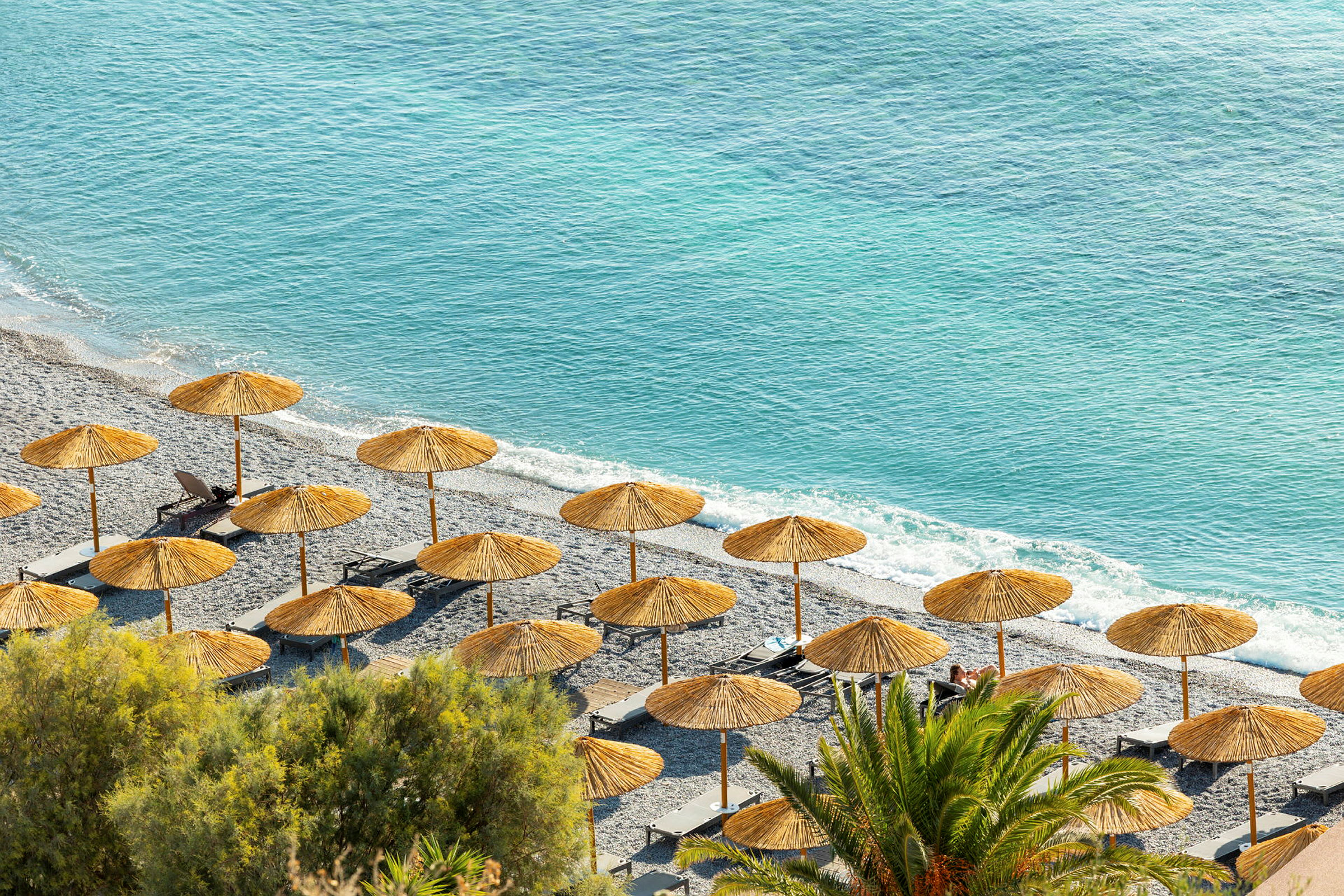 Strand med parasoller og solsenger ved klart blått hav, omgitt av frodig vegetasjon. (AI-generert tekst)