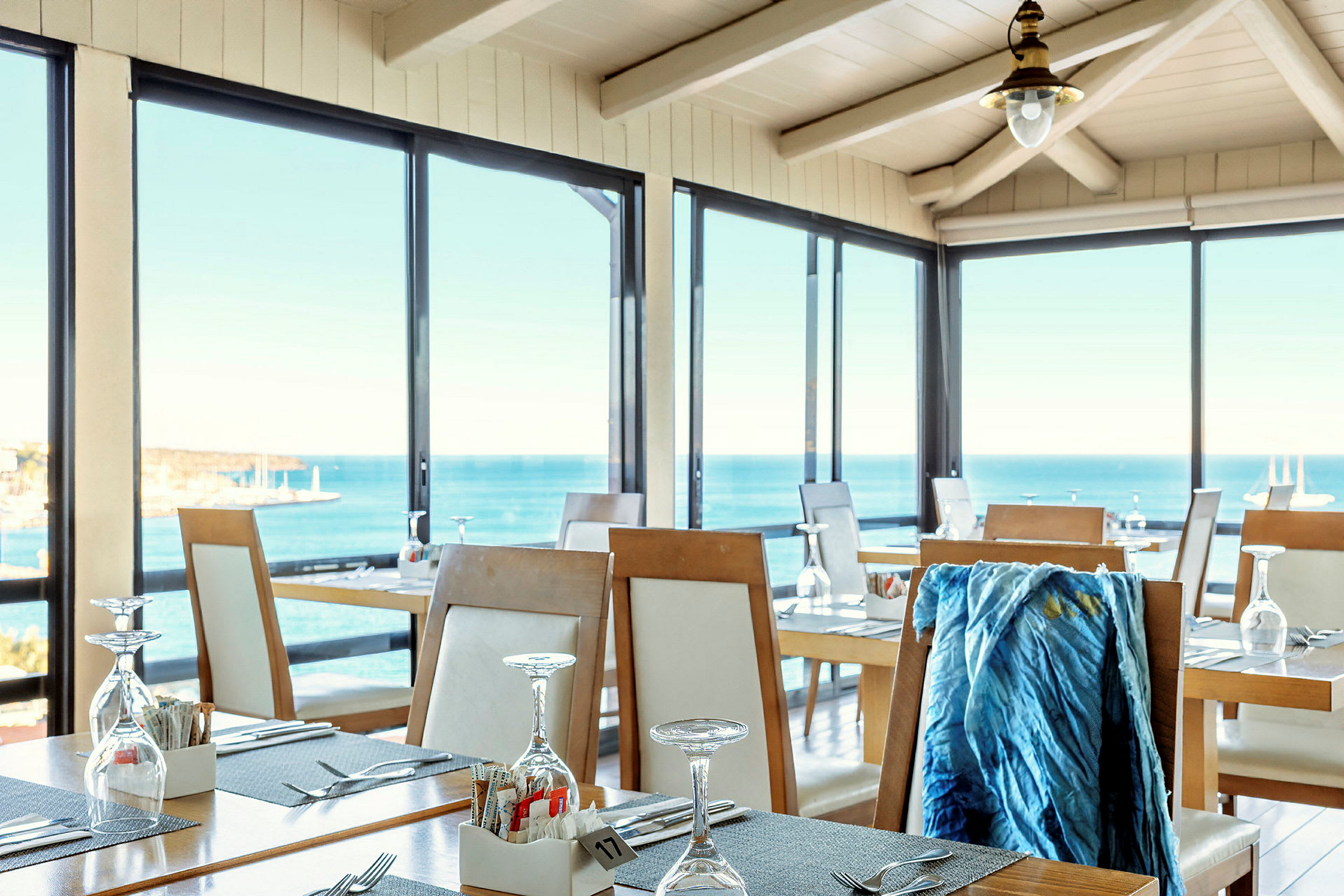 Et lyst og luftig restaurantområde med store glassvinduer som gir utsikt over havet. Bordene er dekket med glass og bestikk. (AI-generert tekst)