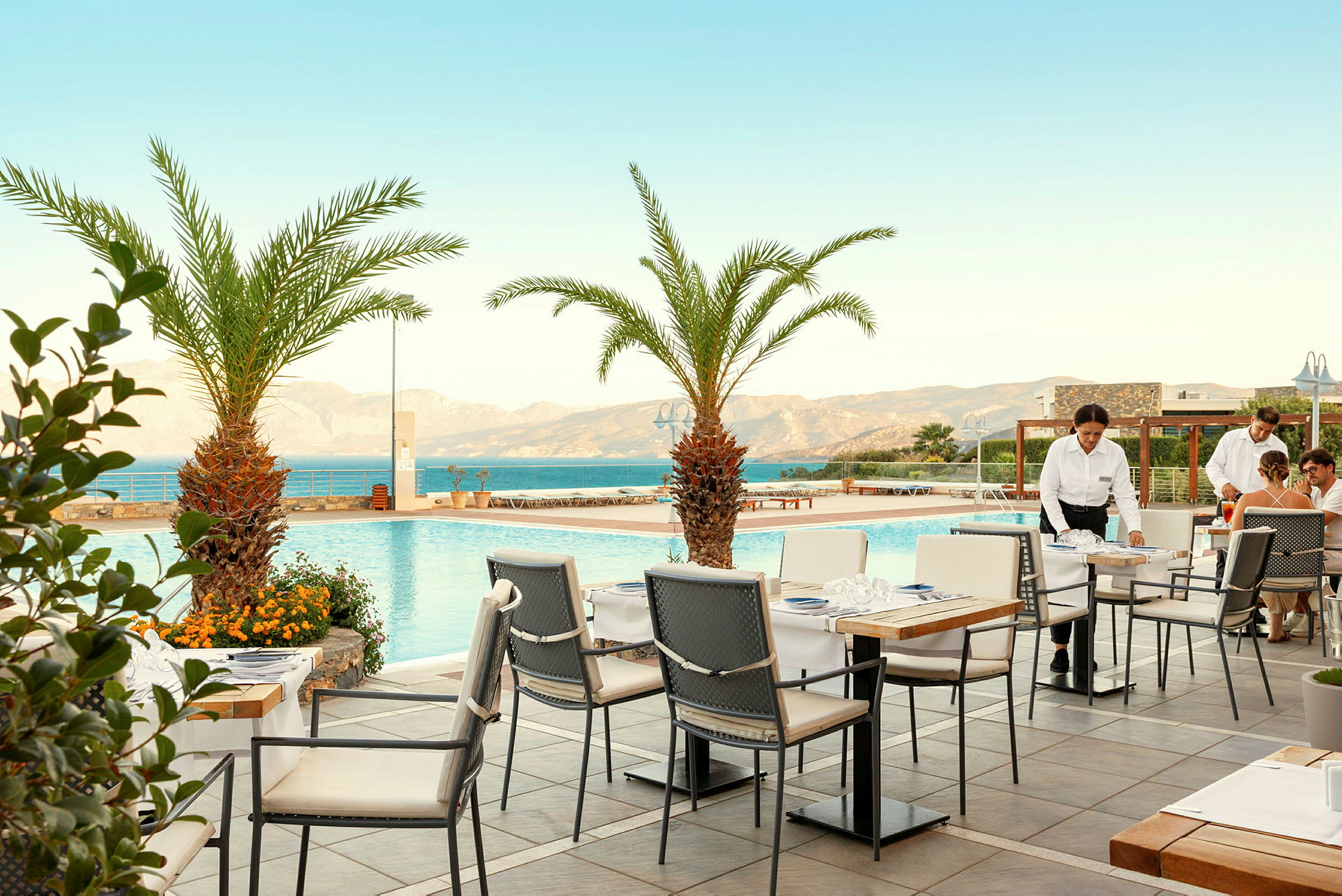En terrasse med utsikt over et basseng og havet, omgitt av palmer. Bord og stoler er plassert på terrassen, og personalet serverer gjester. (AI-generert tekst)