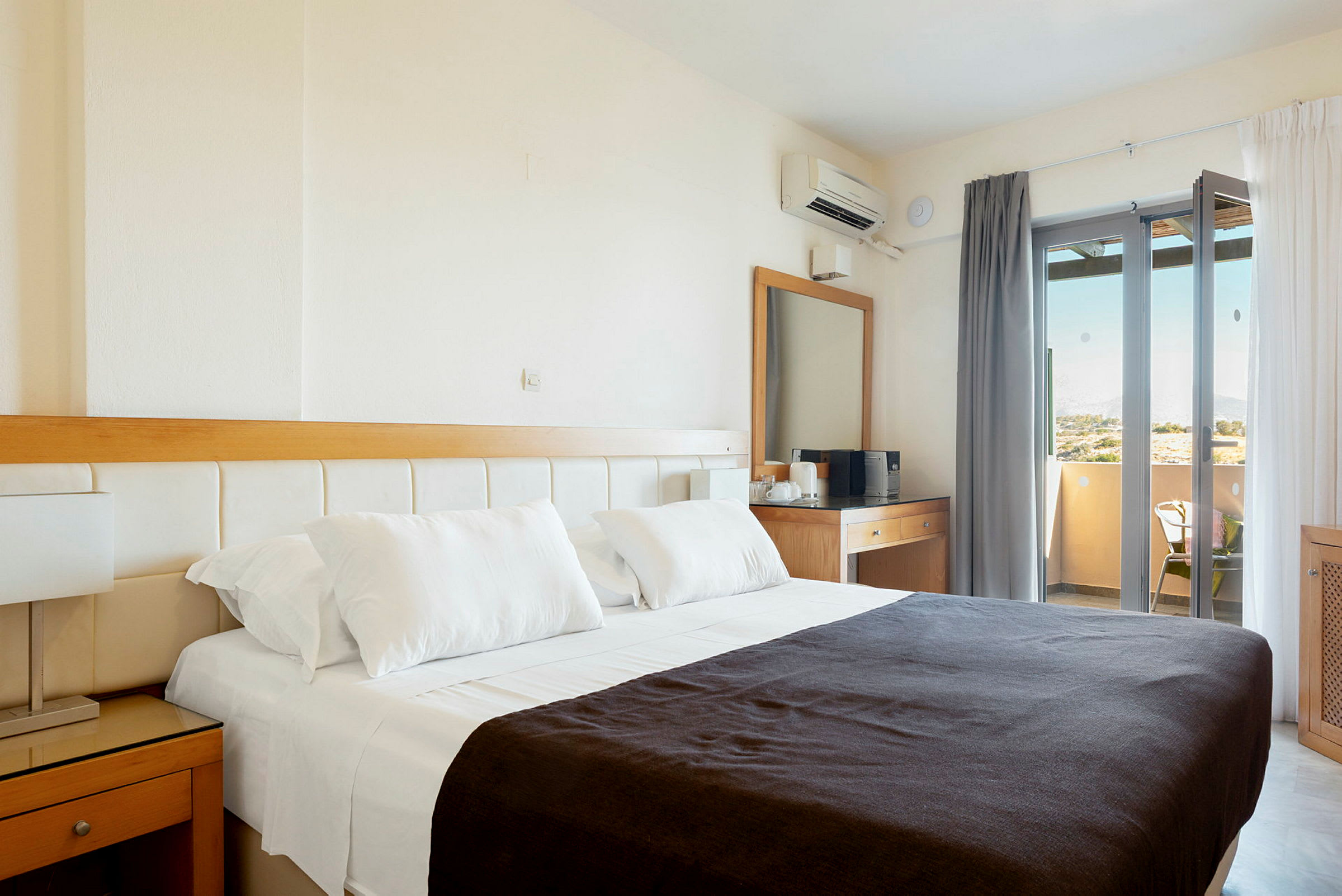 Et hotellrom med en dobbeltseng med hvite laken og et mørkt teppe. Rommet har også et skrivebord med speil og en balkong med utsikt. (AI-generert tekst)