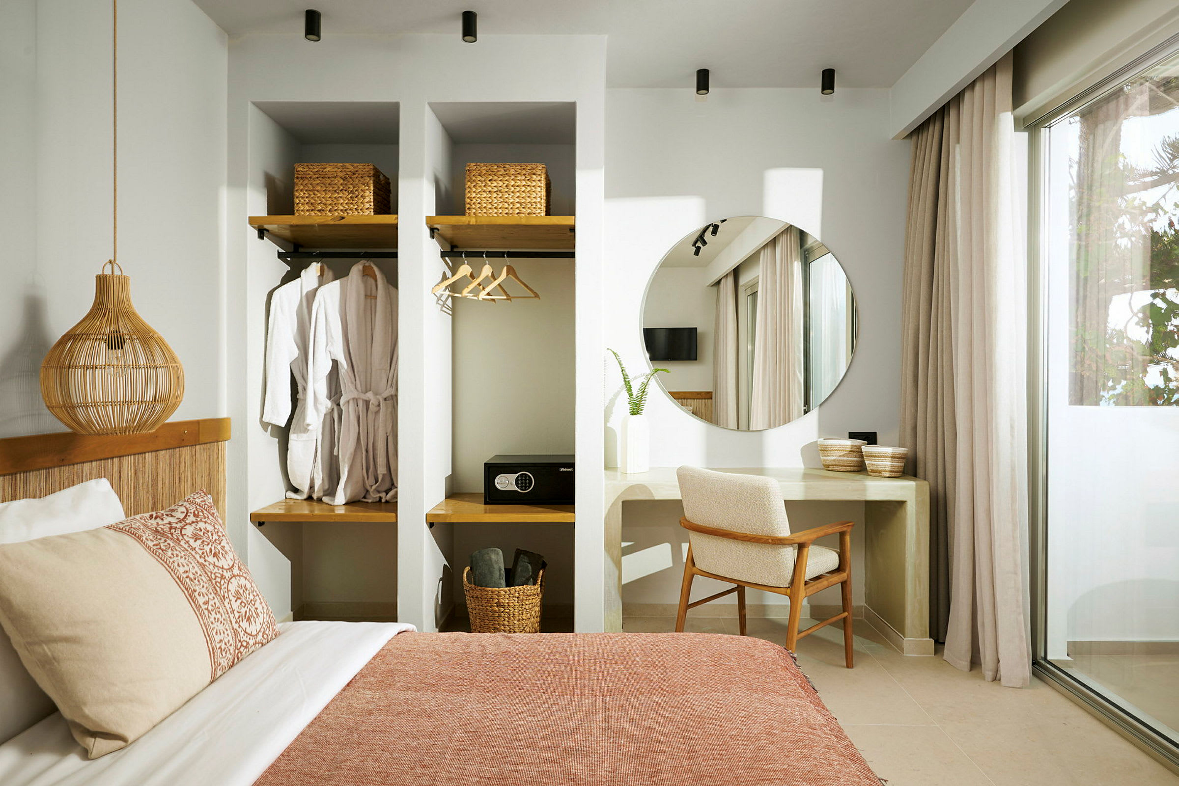 Interiør av et hotellrom med seng, en veggmontert garderobe, et skrivebord med speil og stol, samt store gardiner ved glassdøren. (AI-generert tekst)