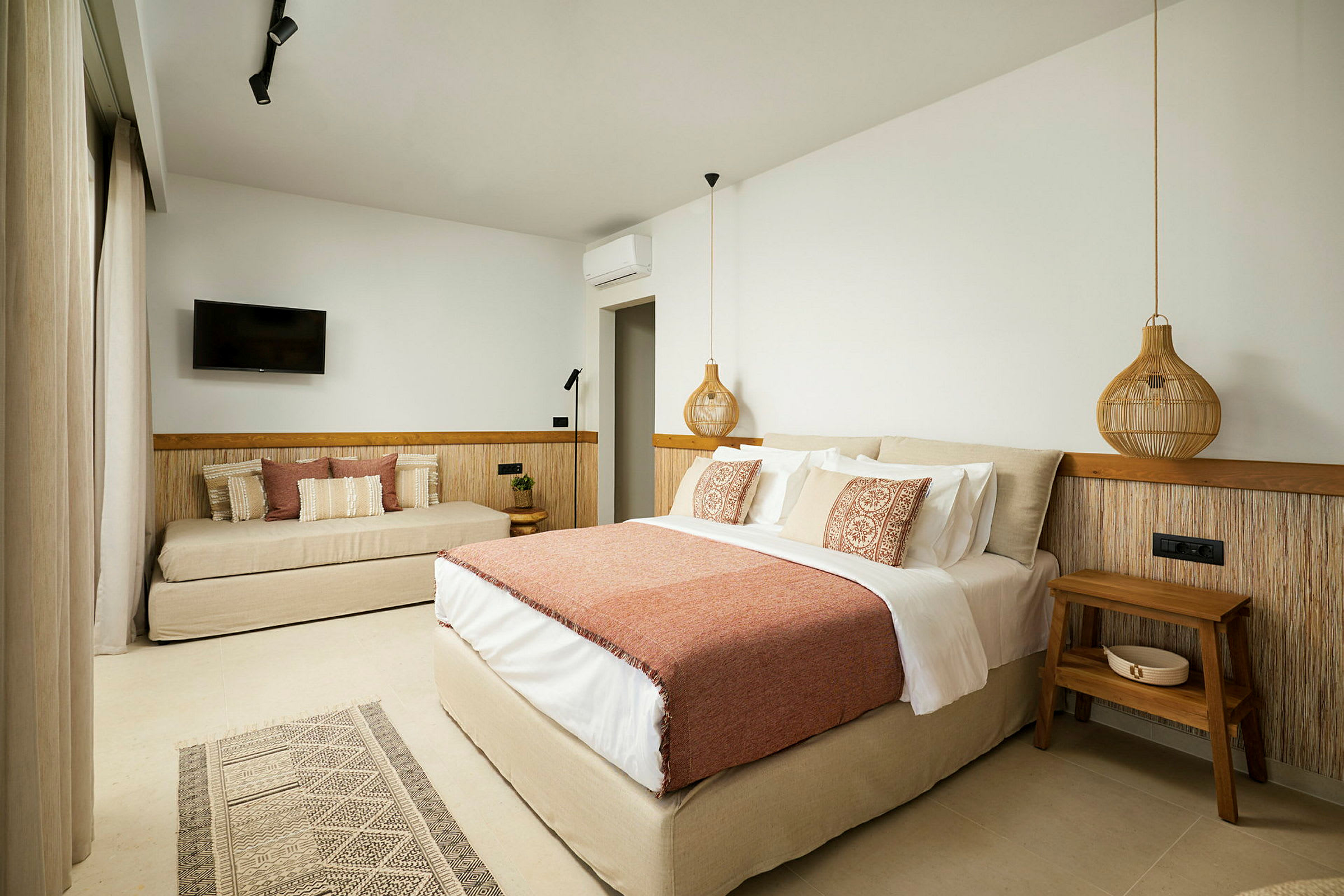 Et hotellrom med en stor seng med oransje teppe og dekorative puter, en sofa med puter, en veggmontert TV og et trebord ved sengen. (AI-generert tekst)