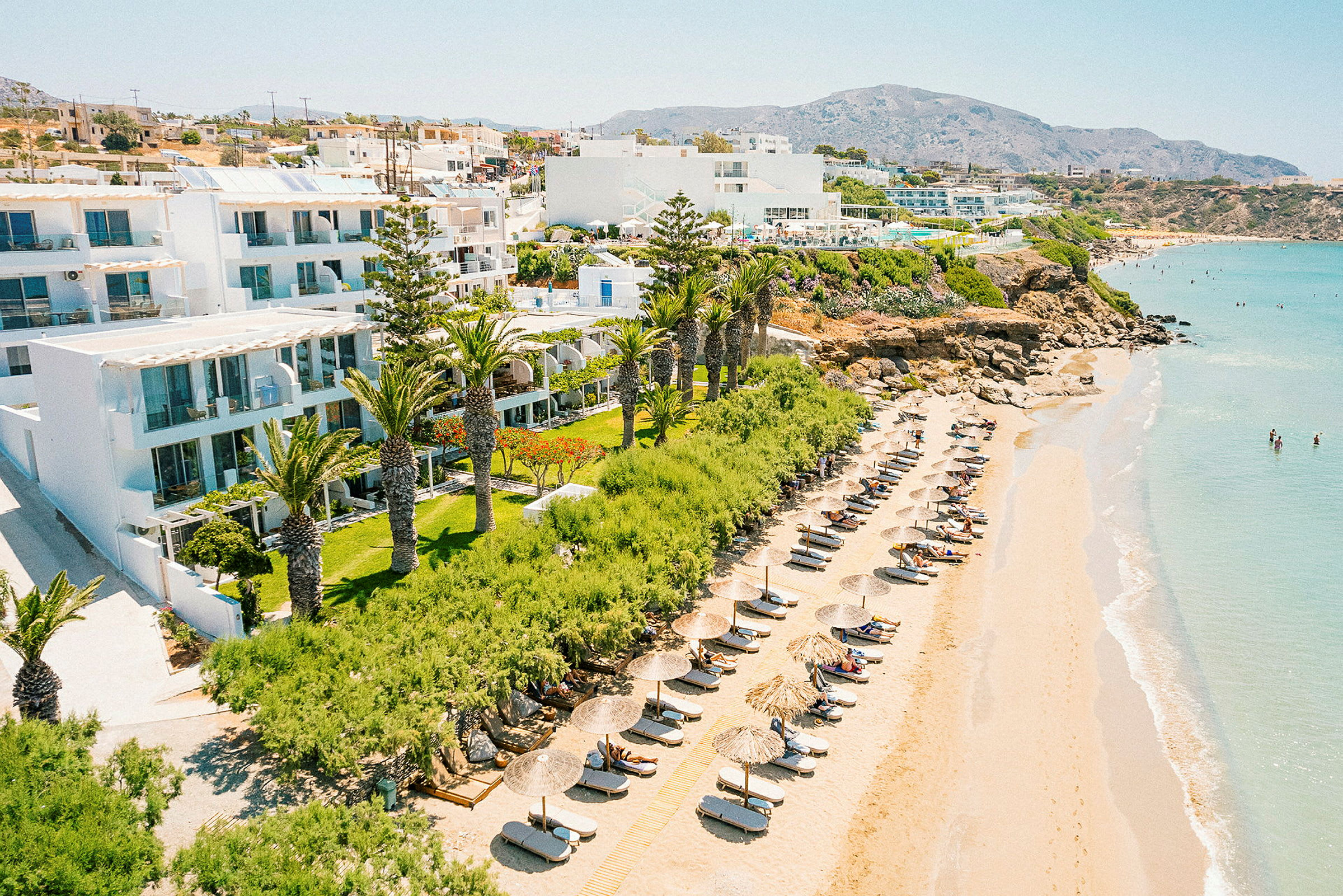 Strand med solsenger og parasoller ved en hotellbygning omgitt av grøntområder. (AI-generert tekst)