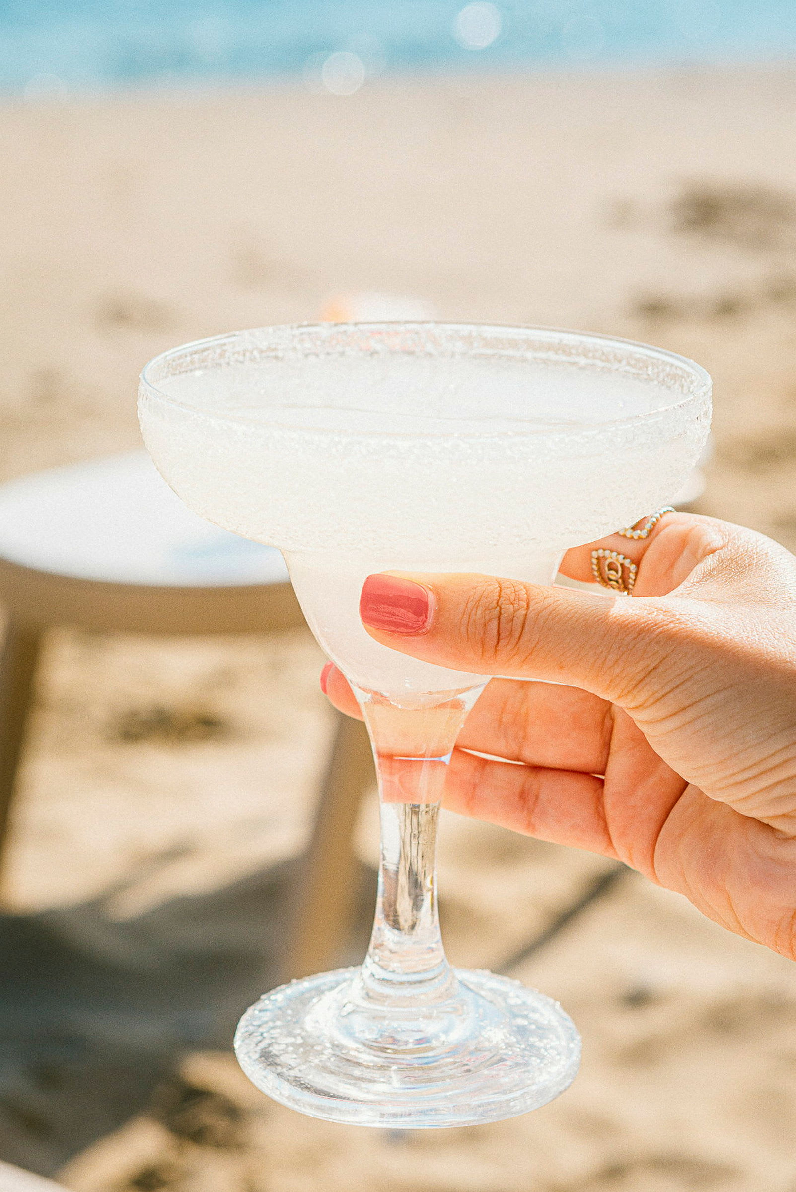 En hånd holder et glass margarita med is ved siden av en strand. (AI-generert tekst)