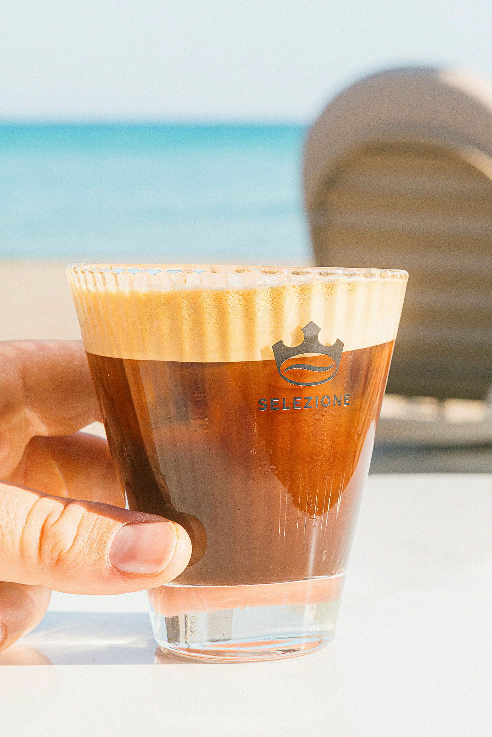 Nærbilde av en hånd som holder et glass med kaffe, stranden er synlig i bakgrunnen. (AI-generert tekst)