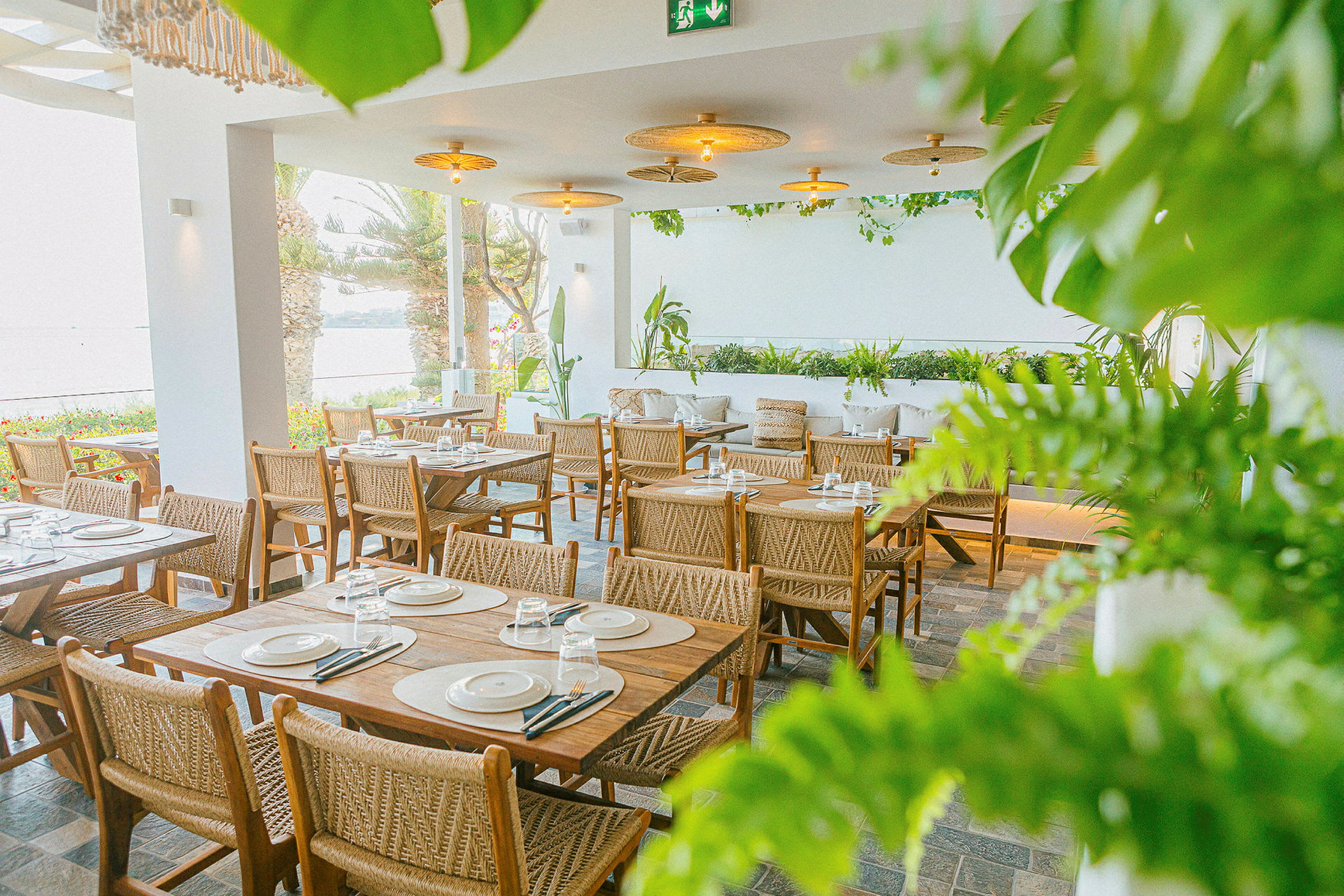 En restaurant med bord og stoler i tre, omgitt av grøntområder og med utsikt over havet. (AI-generert tekst)