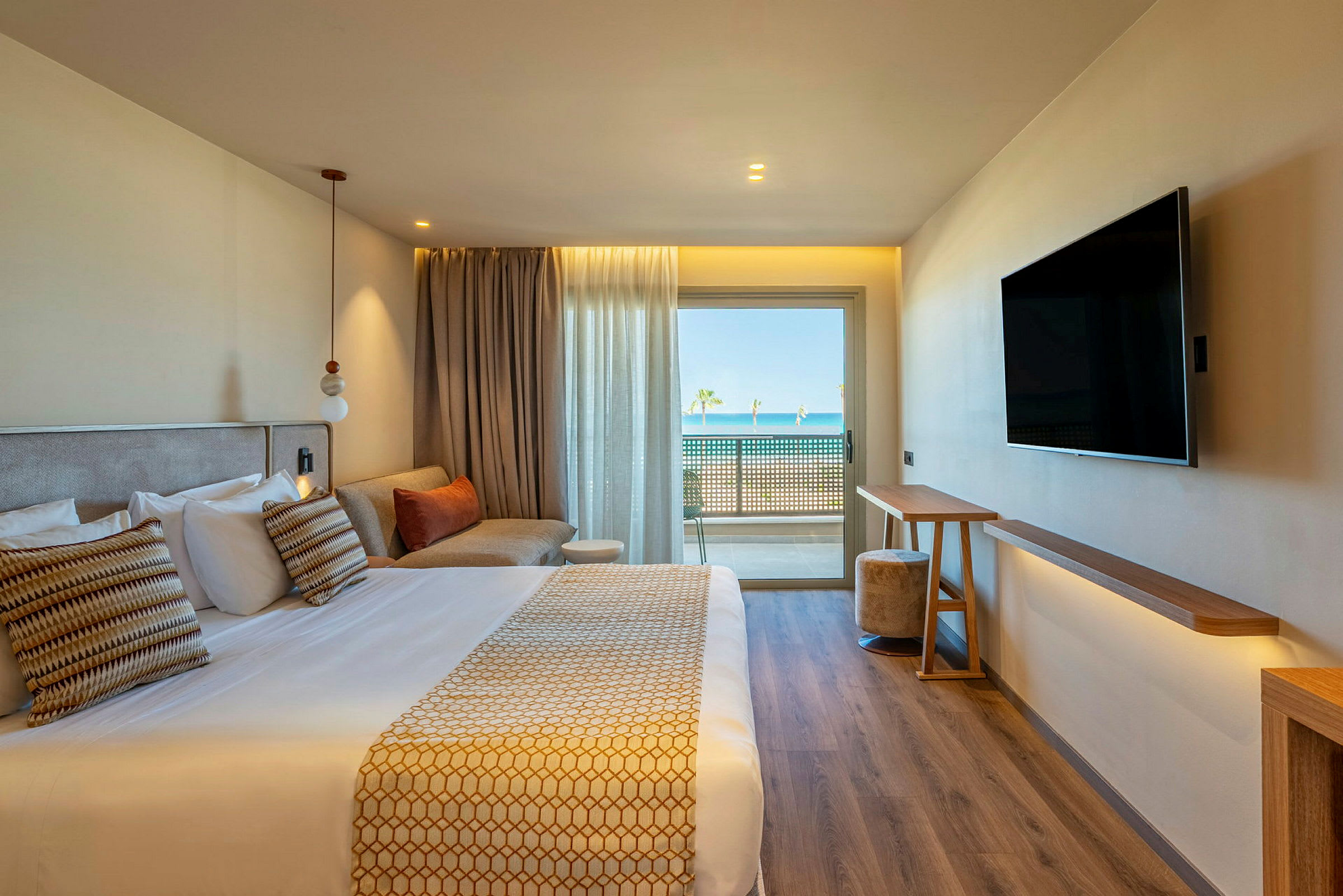 Et moderne hotellrom med en dobbeltseng, en sofa, en veggmontert TV og store vinduer med utsikt mot en balkong og havet i bakgrunnen. (AI-generert tekst)