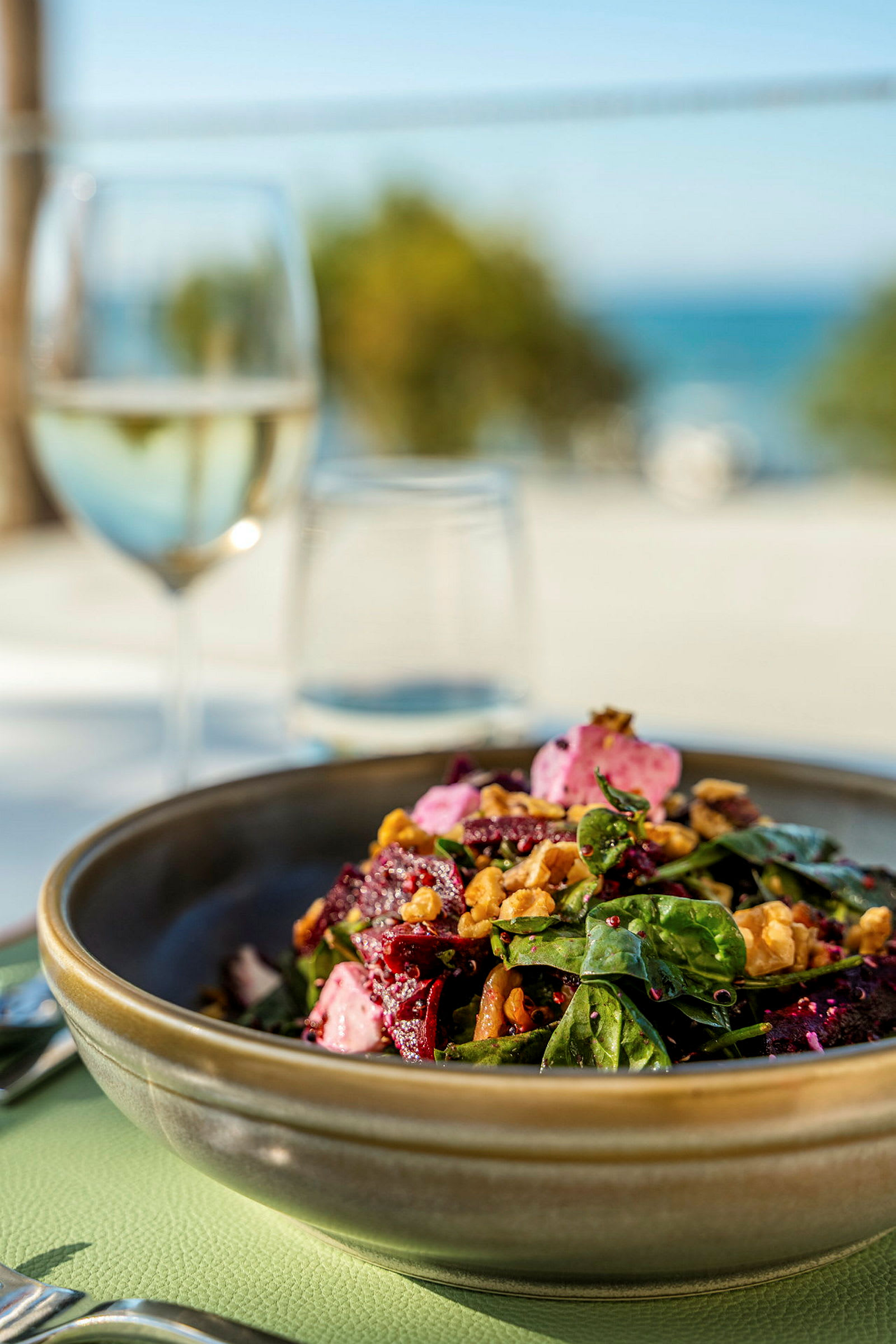 En bolle med salat som inneholder bladgrønnsaker, rødbeter, nøtter og annen topping. I bakgrunnen er det et glass vin og utsikt mot havet. (AI-generert tekst)