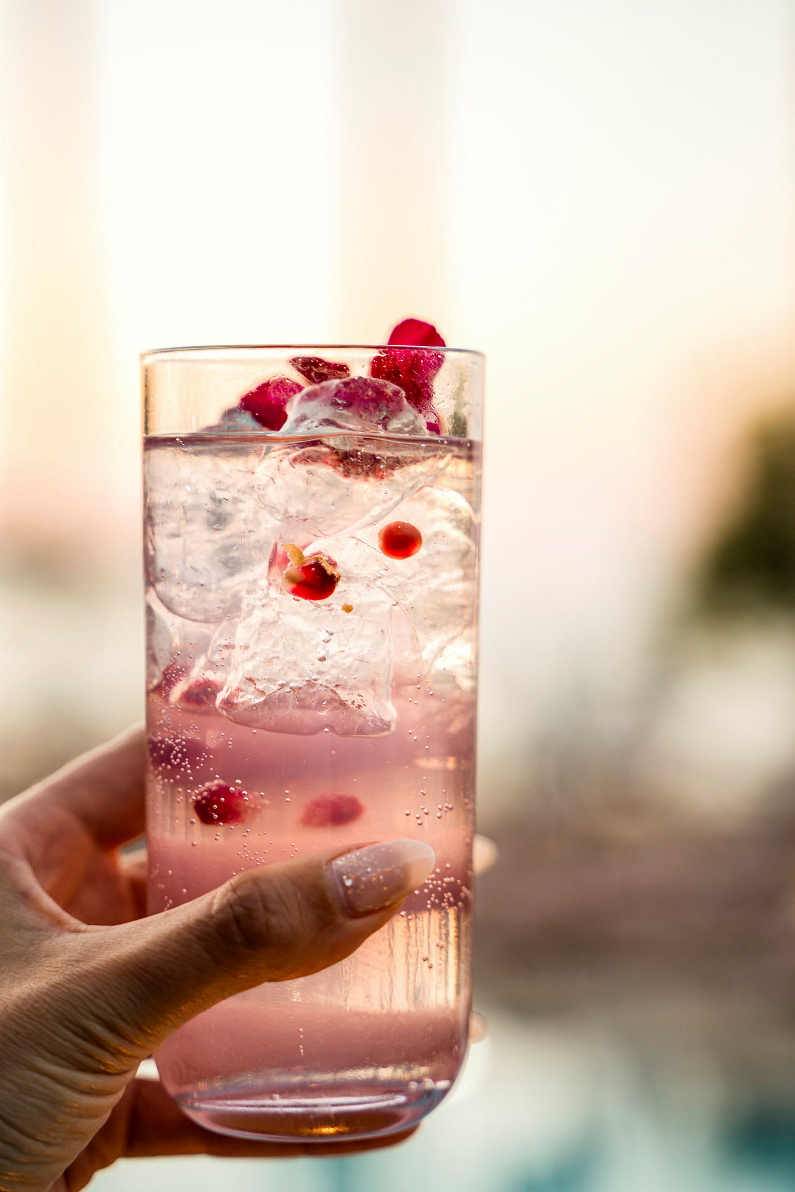 Et glass med rosa drikke, isbiter og granateplekjerner holdes av en hånd. (AI-generert tekst)
