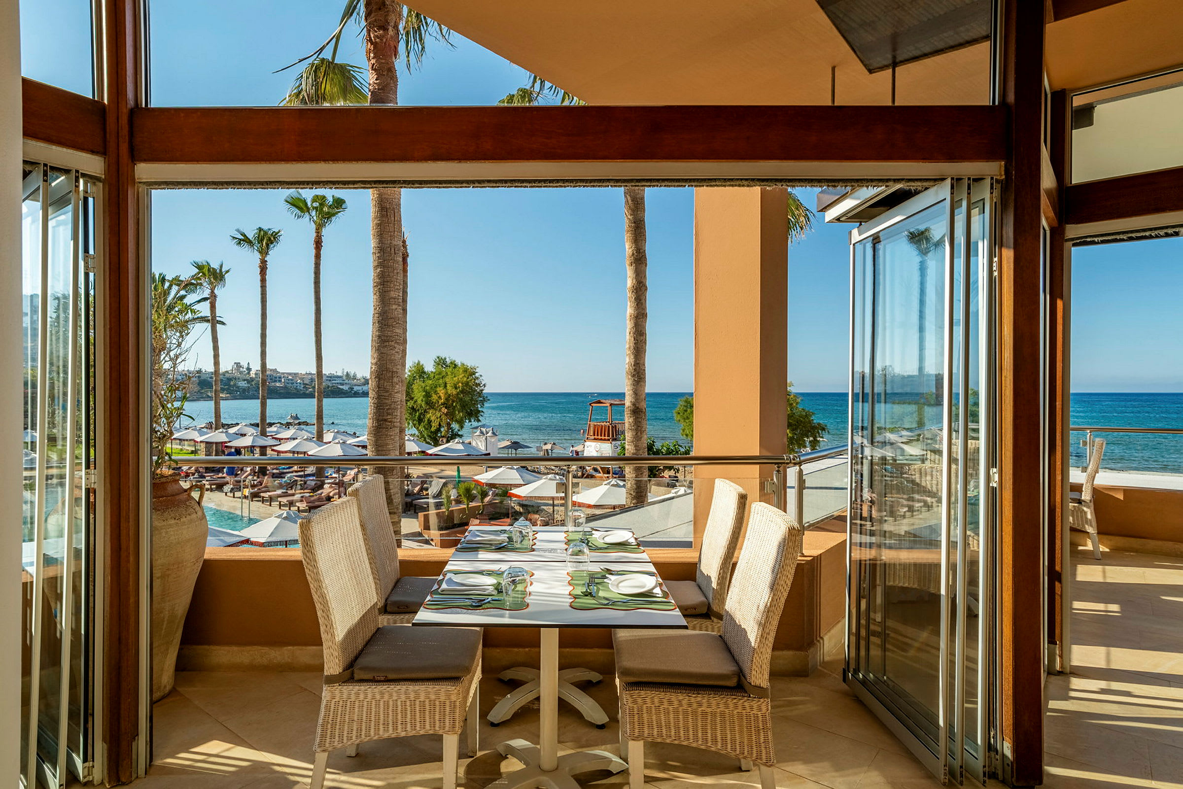 En utendørs restaurant med bord og stoler, utsikt over stranden og havet i bakgrunnen. (AI-generert tekst)