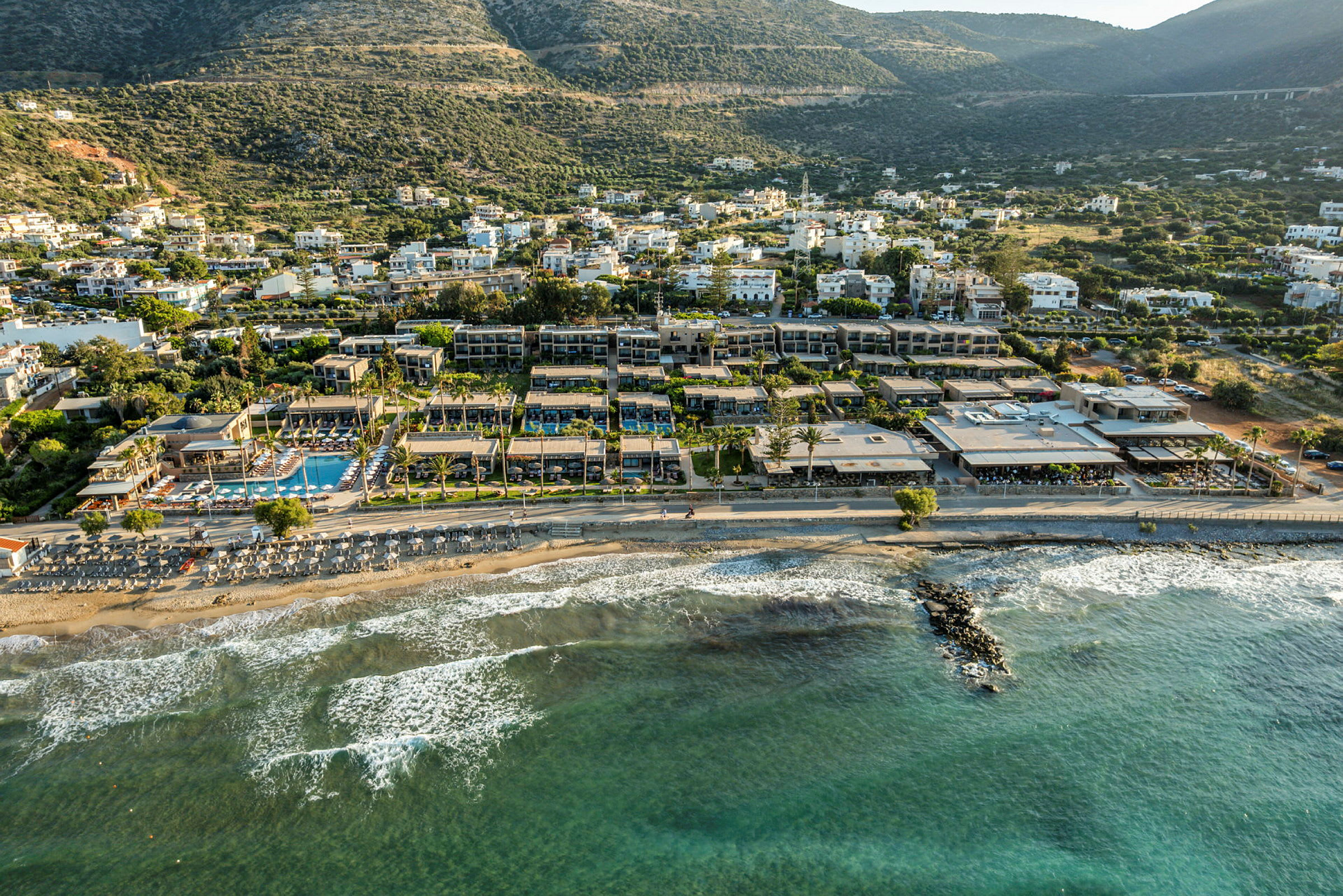 Utsikt over en strand med solsenger ved vannkanten og hotellbygninger i bakgrunnen, med grønne åser lenger bak. (AI-generert tekst)