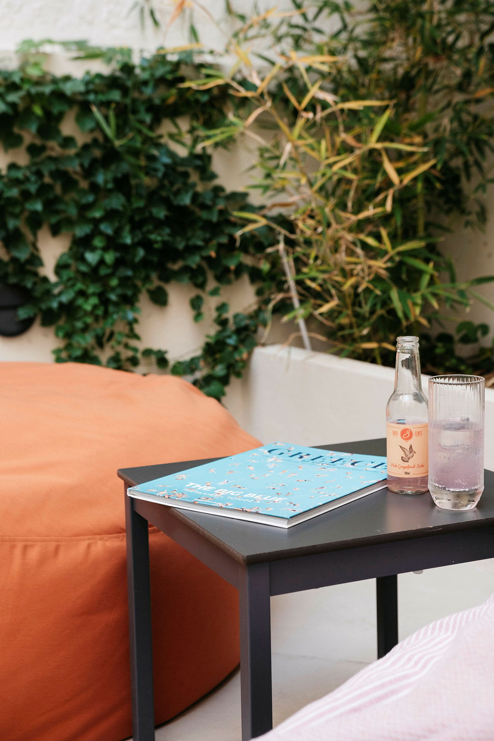 Et bord med et magasin, en flaske og et glass på en uteplass med planter i bakgrunnen. (AI-generert tekst)