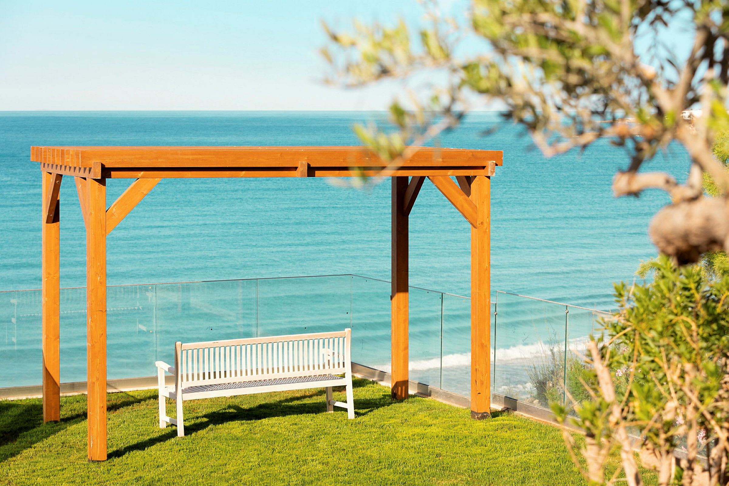 En hvit benk under en pergola av tre på en gressplen med havet i bakgrunnen. (AI-generert tekst)