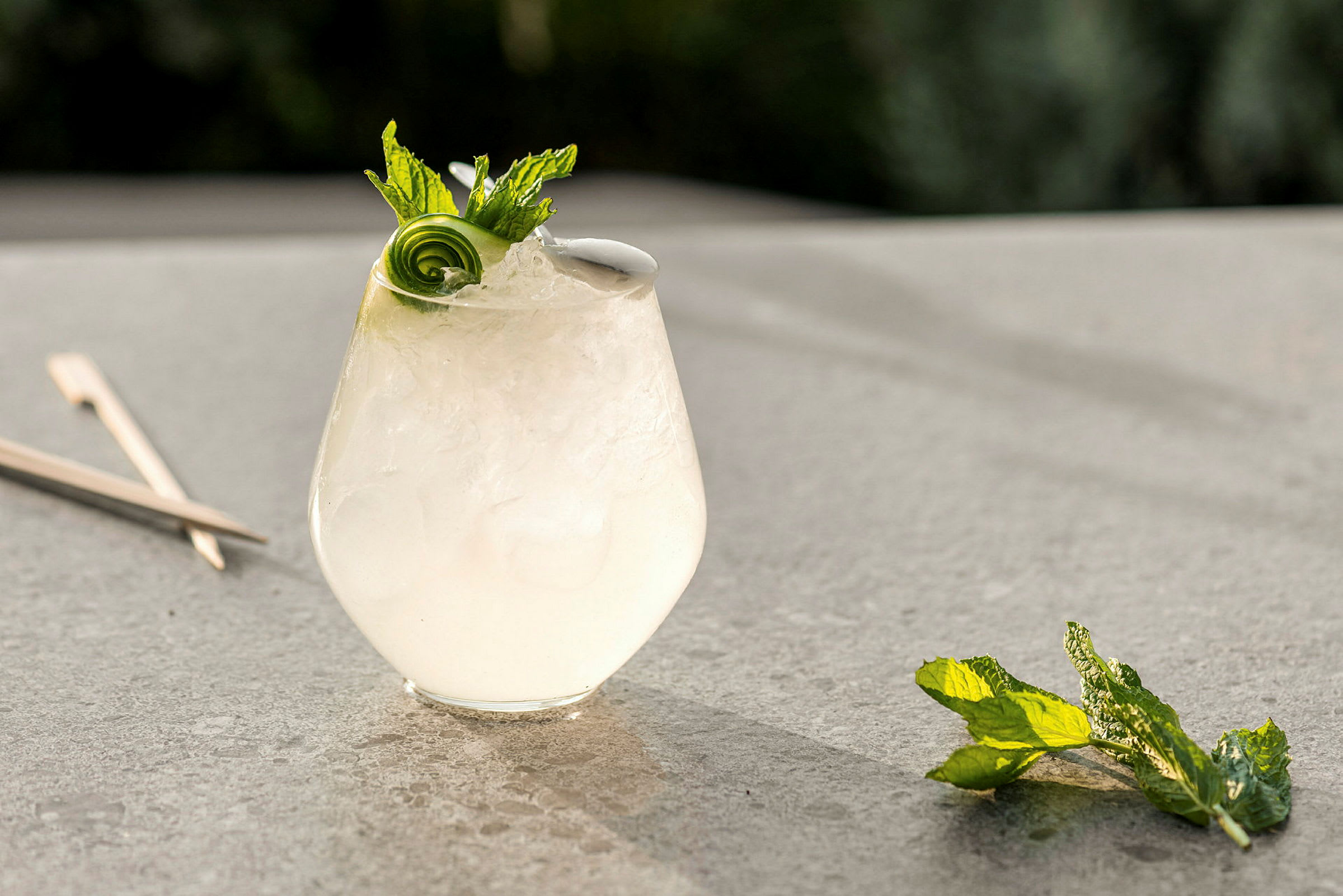 Et glass med en kald drink, pyntet med mynte og agurkspiral, står på en bordflate. (AI-generert tekst)