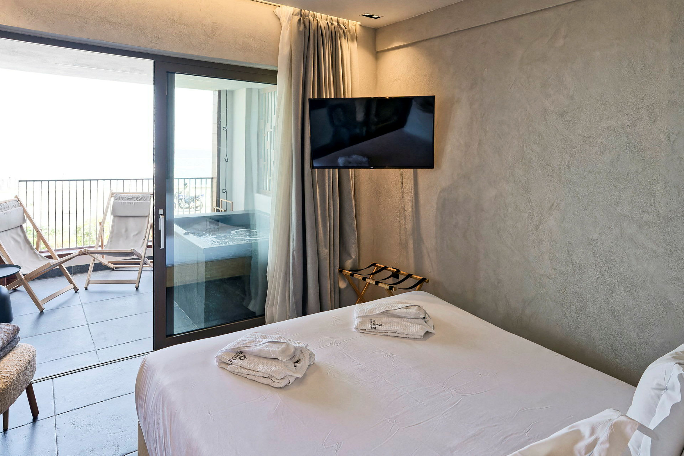 Moderne hotellrom med hvit dobbeltseng som åpner ut til en balkong med solsenger og boblebad. (AI-generert tekst)