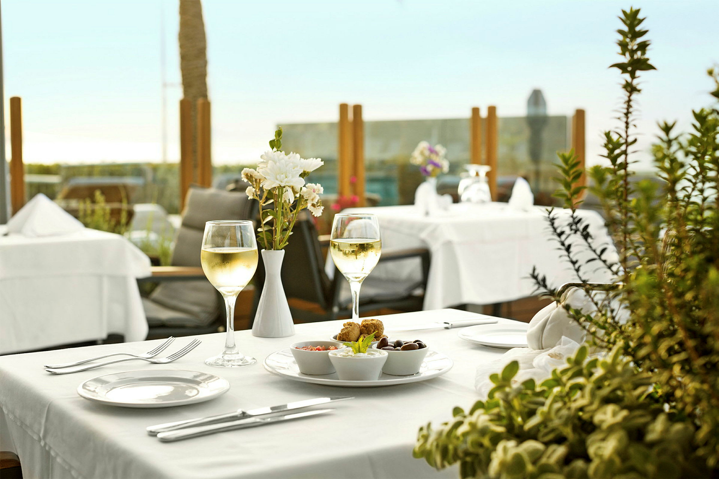 Et dekket restaurantbord med et glass hvitvin, blomsterdekorasjon og tallerken med snacks. (AI-generert tekst)
