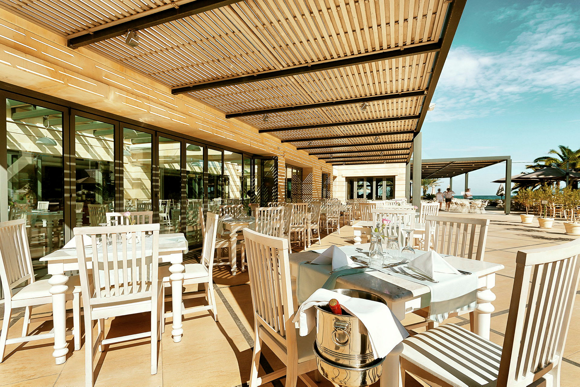 Utendørs restaurant med hvite bord og stoler samt pergola som gir skygge. (AI-generert tekst)