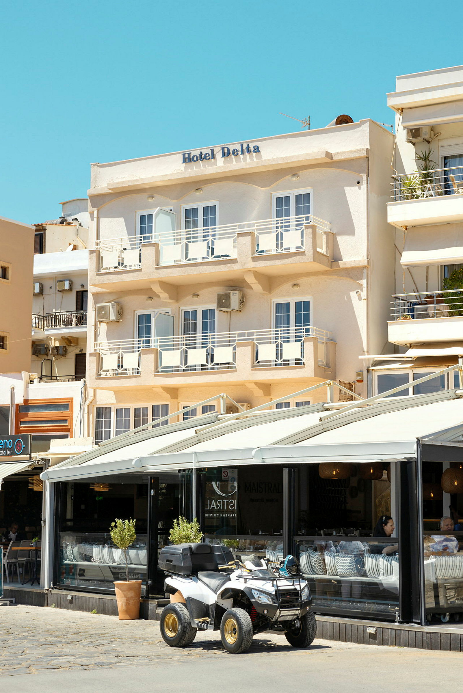Fasaden til et hotell med navnet "Hotel Delta" med balkonger og en kafe i første etasje, hvor en firehjuling er parkert utenfor. (AI-generert tekst)