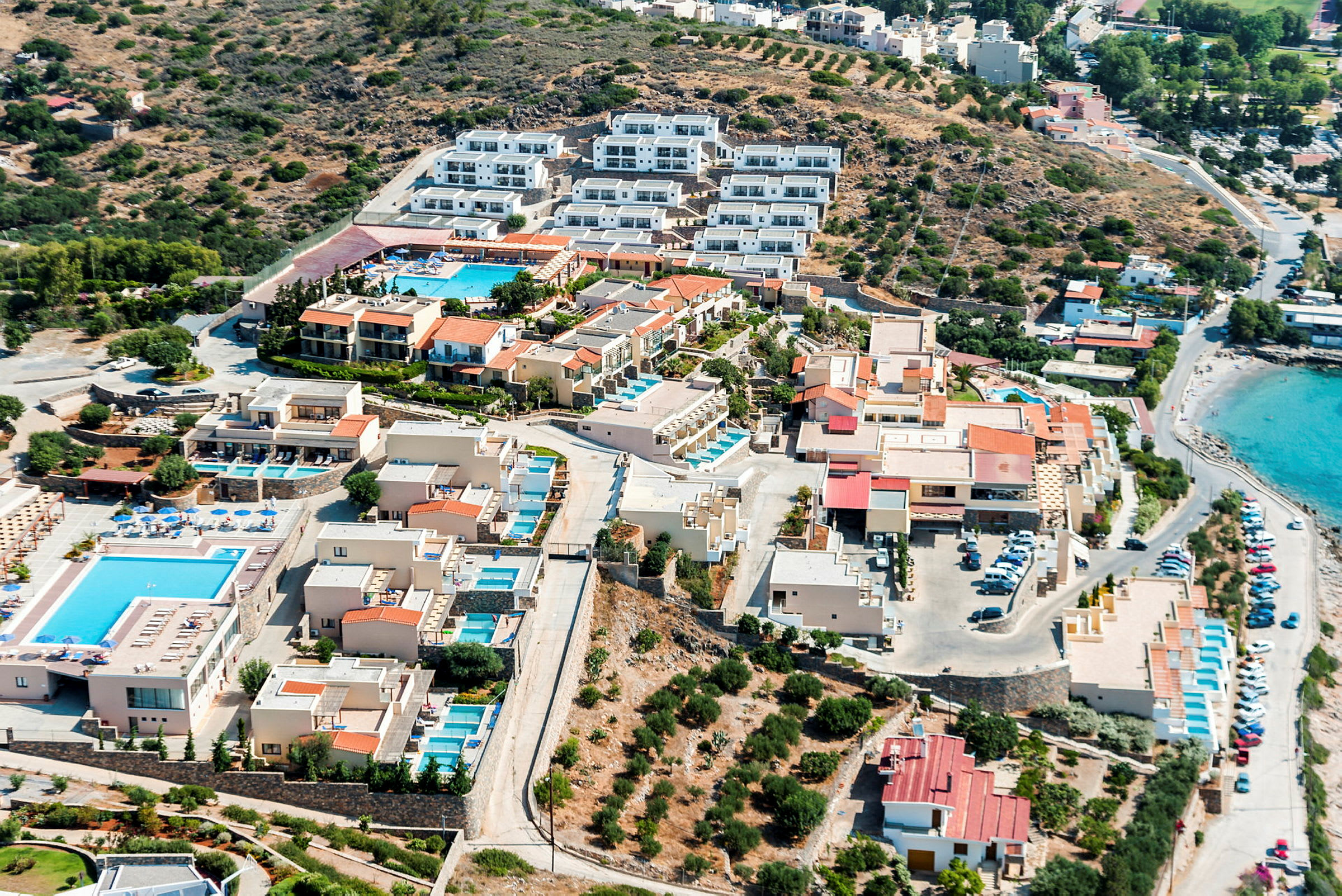 Flyfoto av hotellområde med bassenger og strand. (AI-generert tekst)