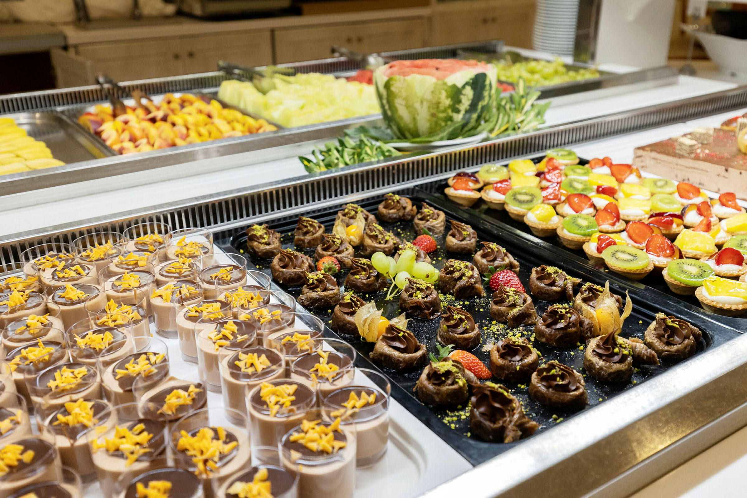 Buffet med forskjellige desserter, inkludert sjokolademousse, fruktdekorasjoner og små kaker. (AI-generert tekst)