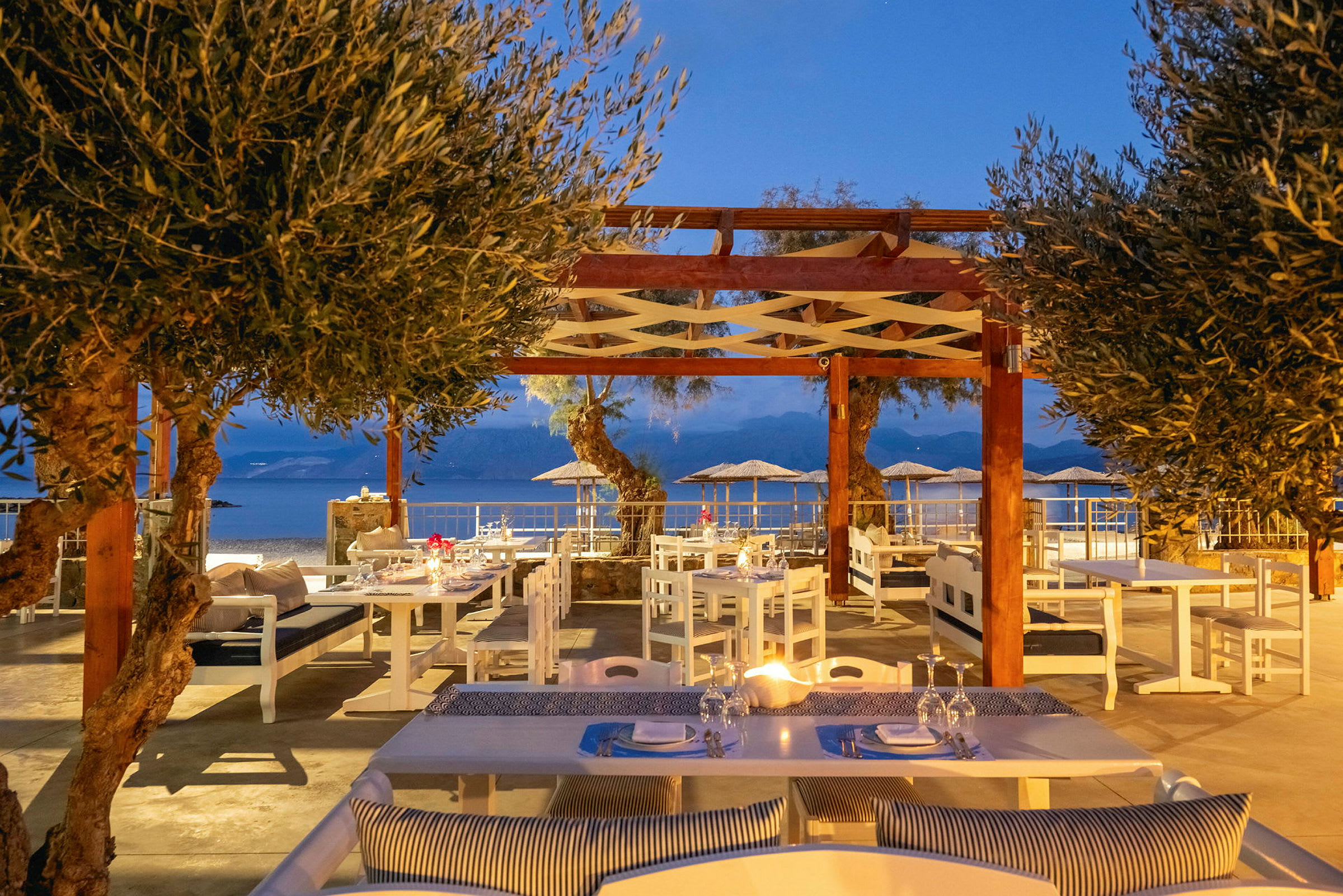 En utendørsrestaurant med sitteplasser under en trepergola og utsikt mot havet. (AI-generert tekst)
