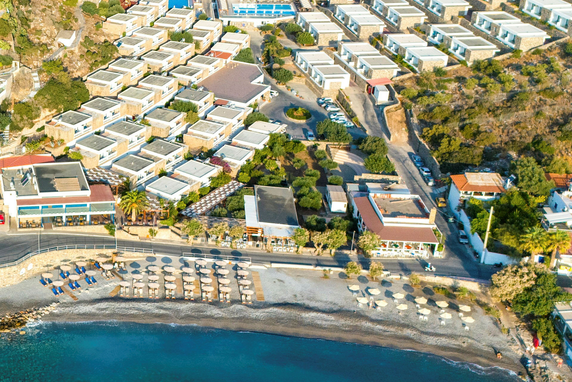 Luftfoto av et hotellområde med basseng, bilparkering og nærliggende strand med parasoller. (AI-generert tekst)